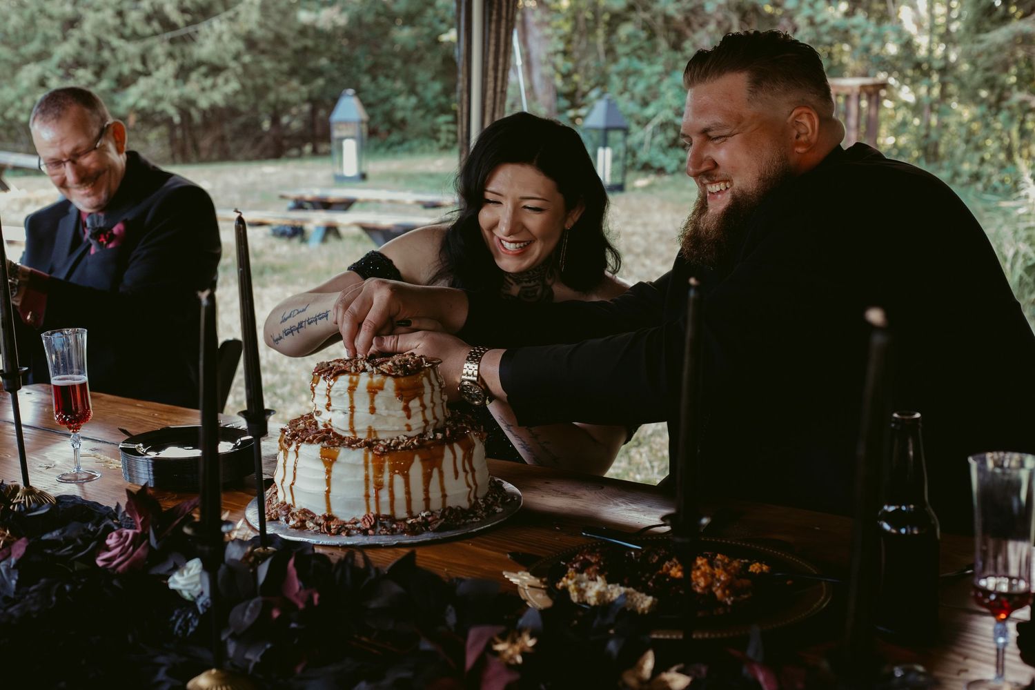 Couple cutting caramel drip wedding cake together at outdoor rustic reception table with guests nearby.