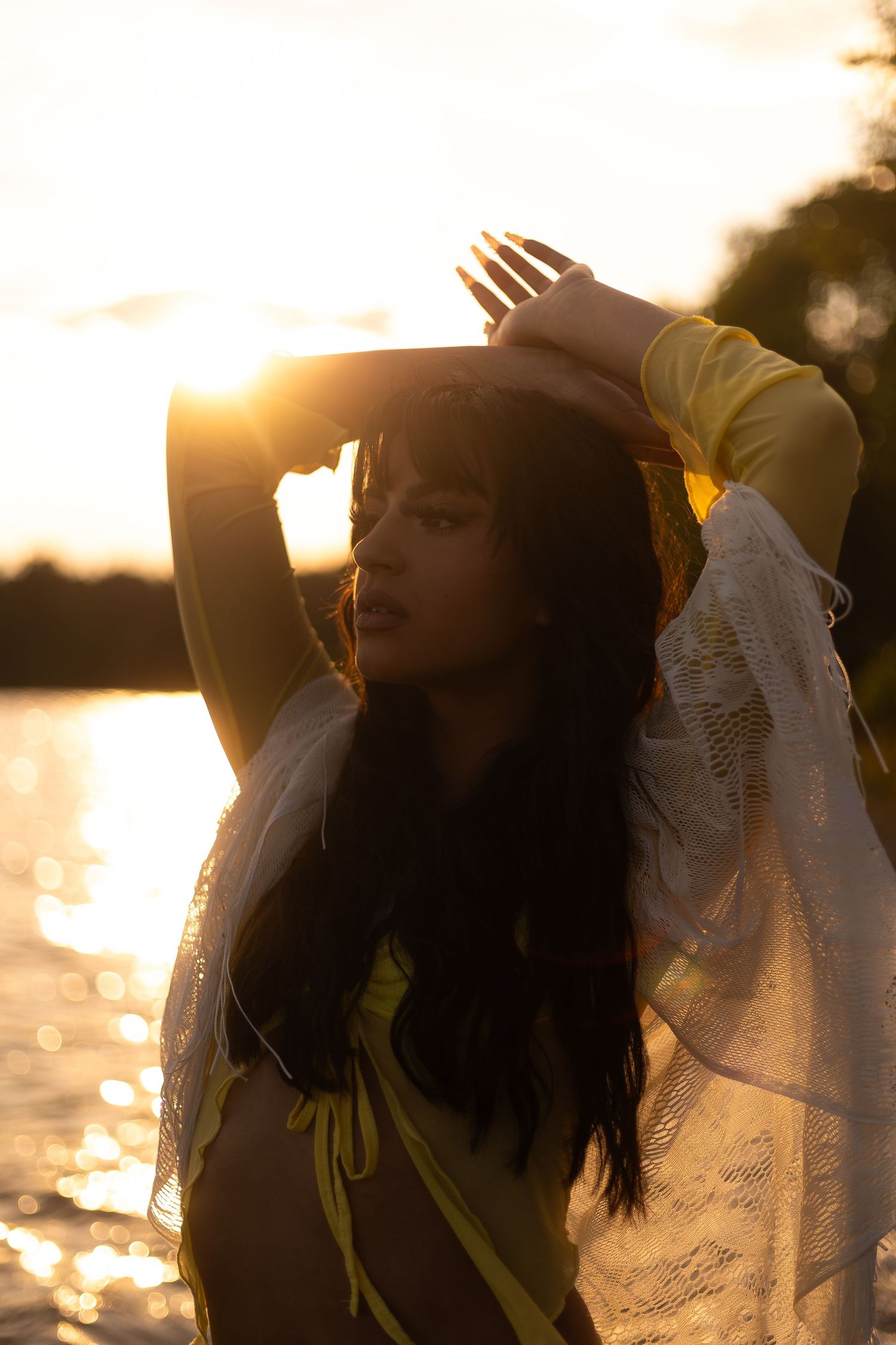 A woman poses gracefully at sunset by the water wearing a yellow bikini and sheer wrap.