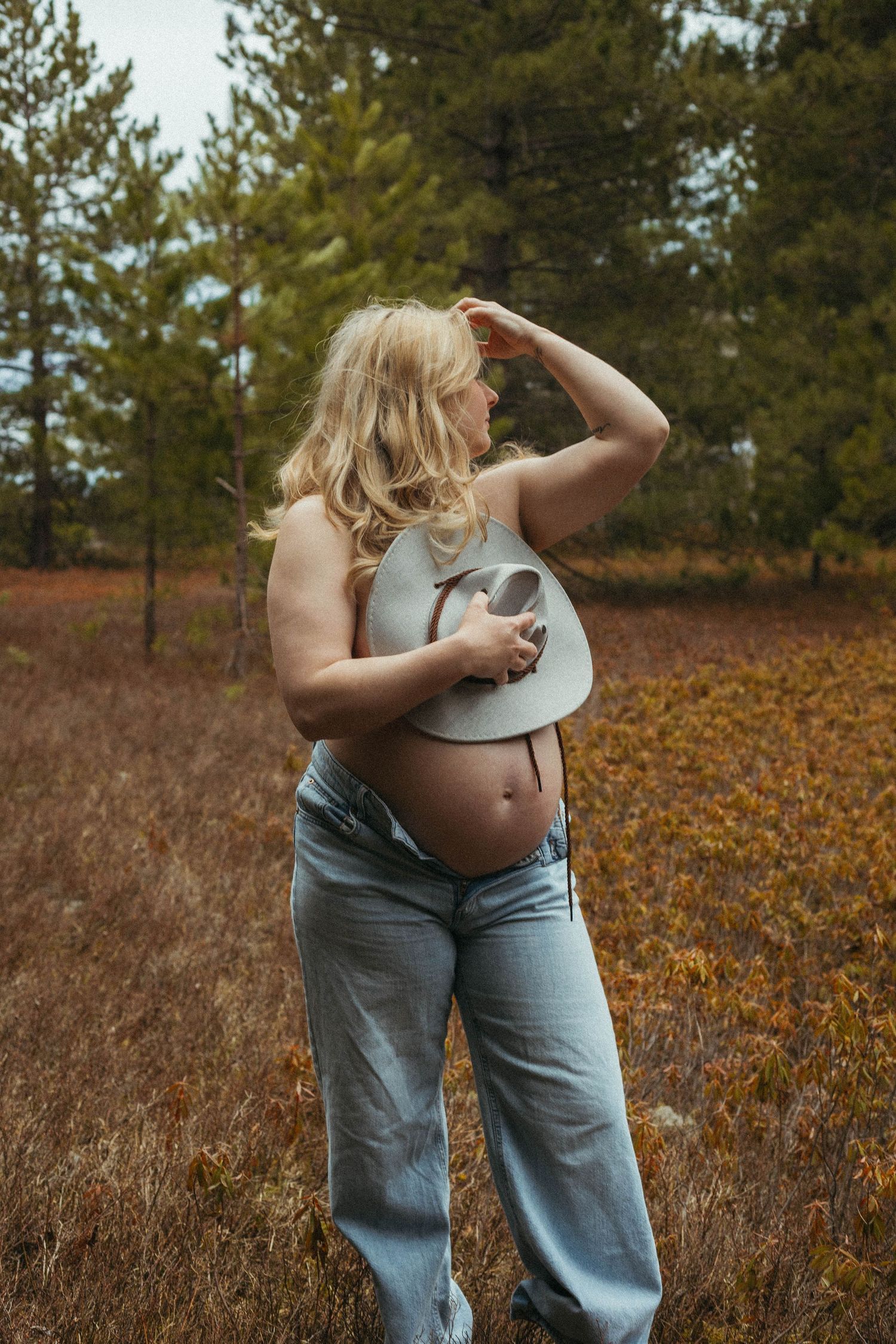 Pregnant woman in jeans holding cowboy hat in autumn forest field, showing baby bump outdoors.