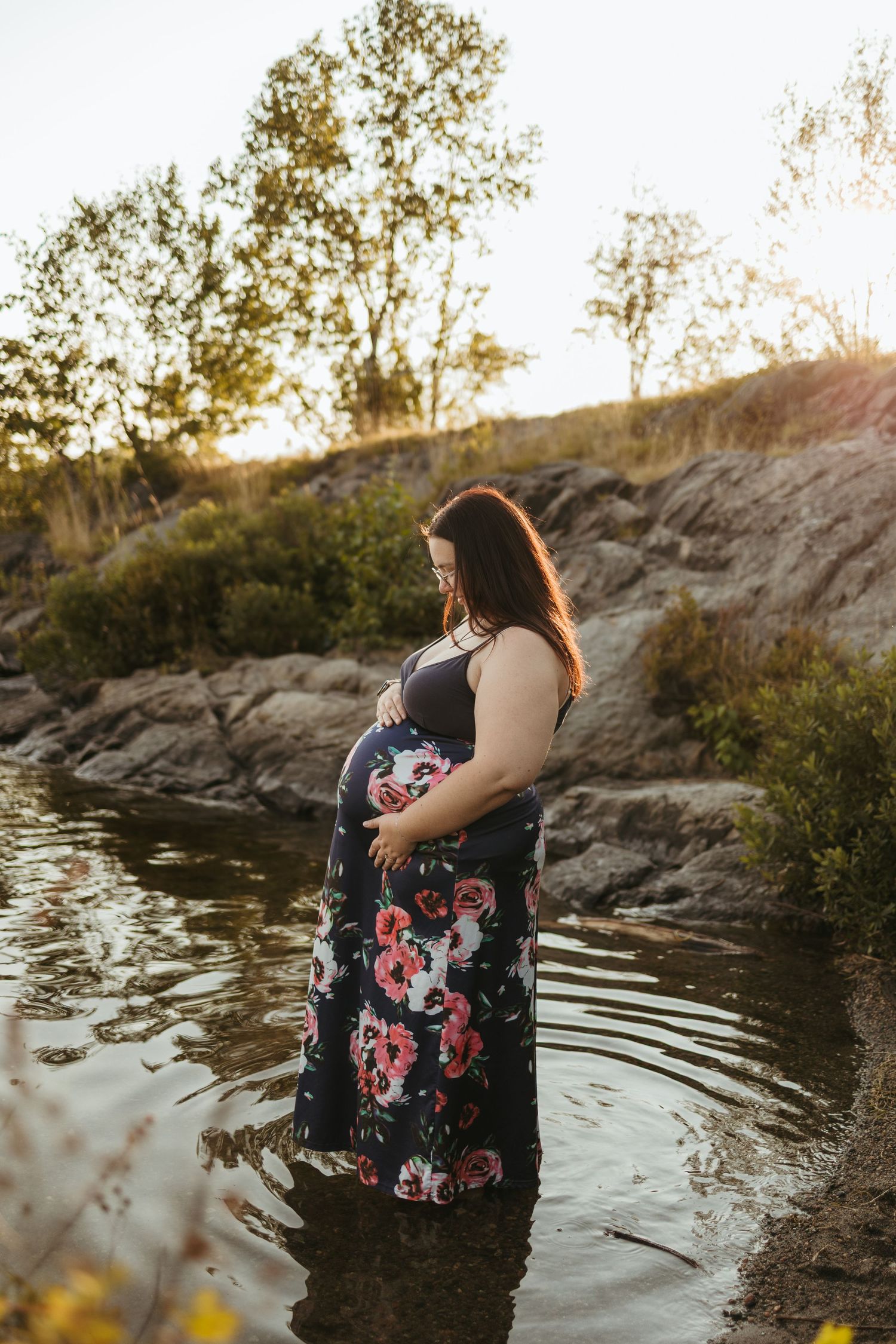 Pregnant woman in floral dress standing in shallow water at golden hour, cradling her baby bump outdoors.
