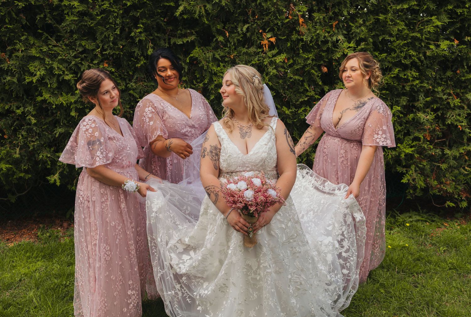 Bride in white lace gown holds pink bouquet surrounded by three bridesmaids in dusty rose dresses outdoors.