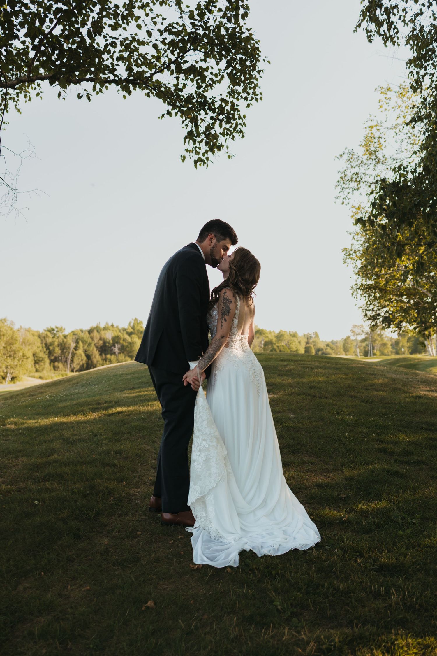 Bride and groom share a romantic forehead touch on a lush green hillside, framed by trees on a sunny day.
