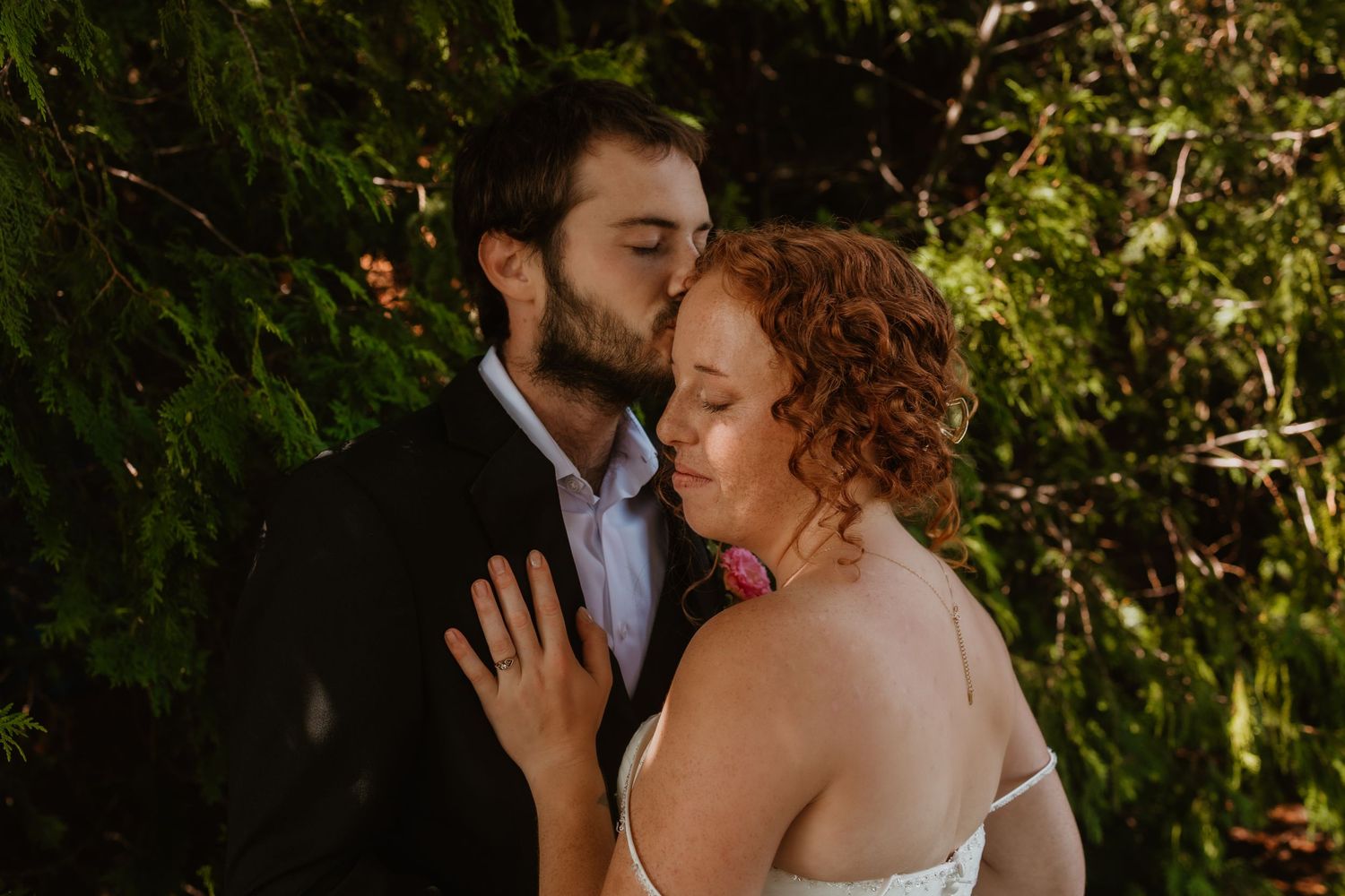 Groom kissing bride on cheek in romantic outdoor wedding portrait surrounded by lush green foliage.