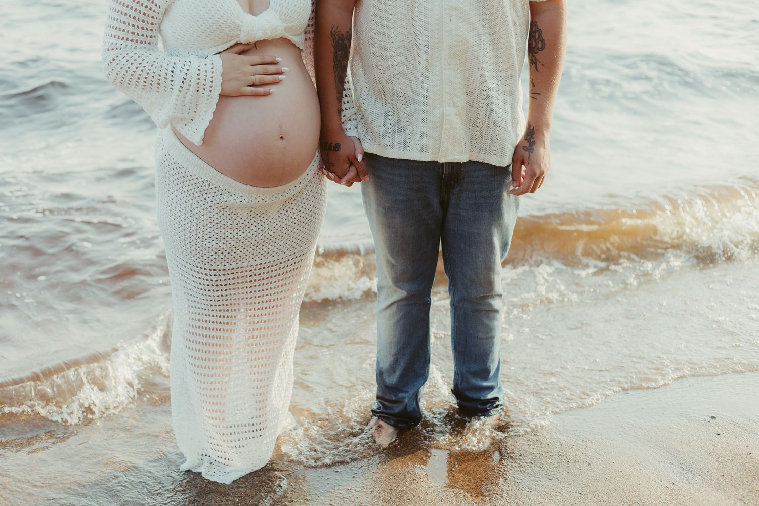 Pregnant woman in white crochet dress holding hands with partner while standing in ocean waves.