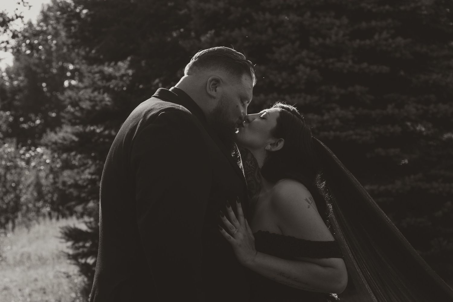 Black and white photo of couple sharing a romantic kiss outdoors surrounded by trees.