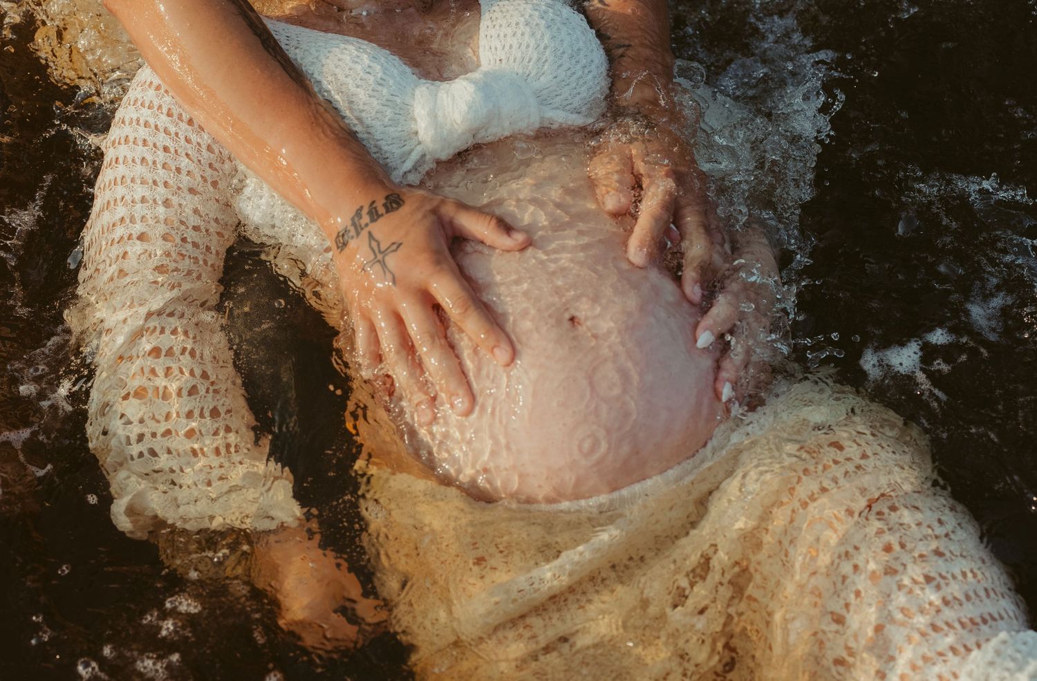 Close-up of pregnant belly in white crochet dress splashed with water.