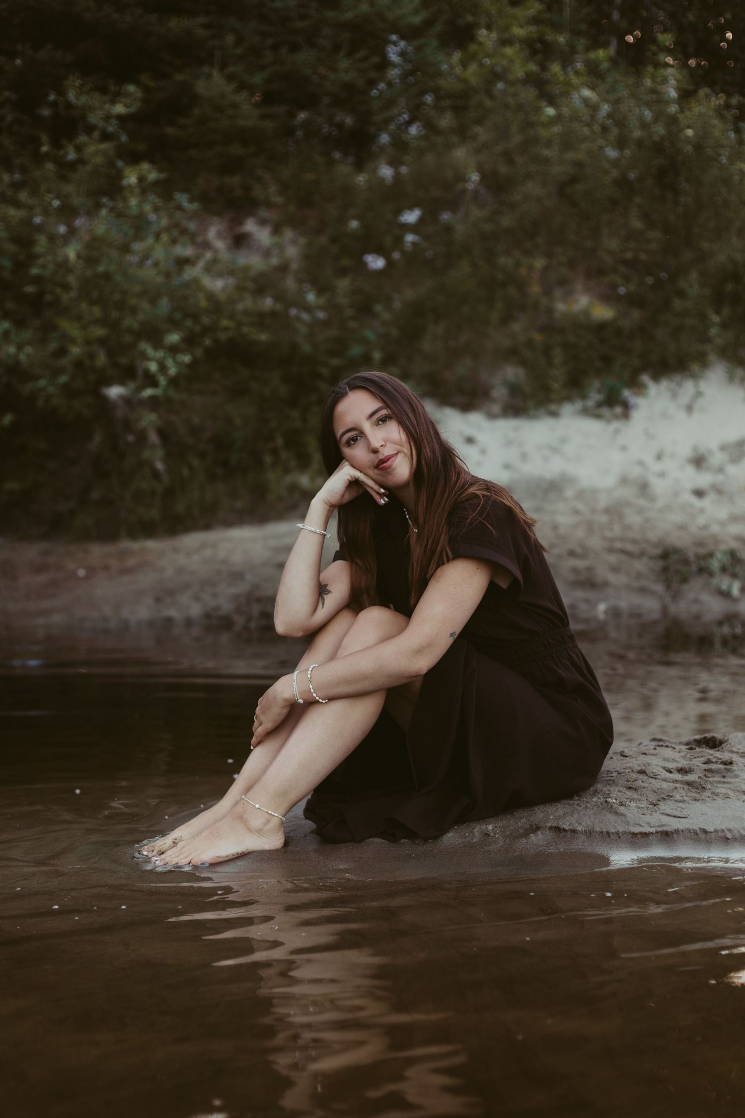 Woman in black dress sitting barefoot by water's edge with lush green trees in background.