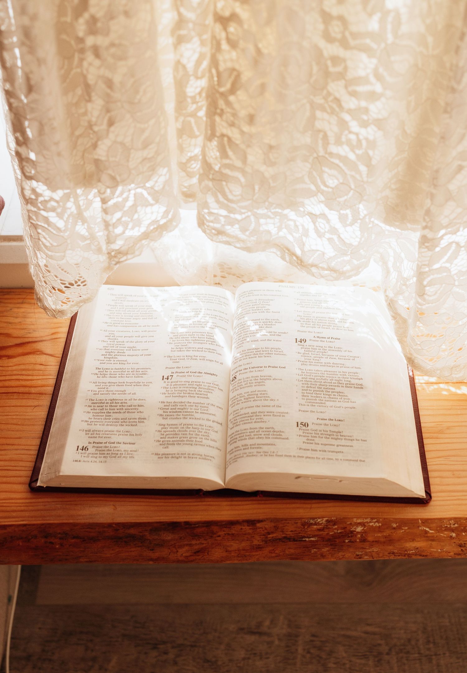Open Bible resting on a wooden bench in front of a white lace curtain in soft natural light.