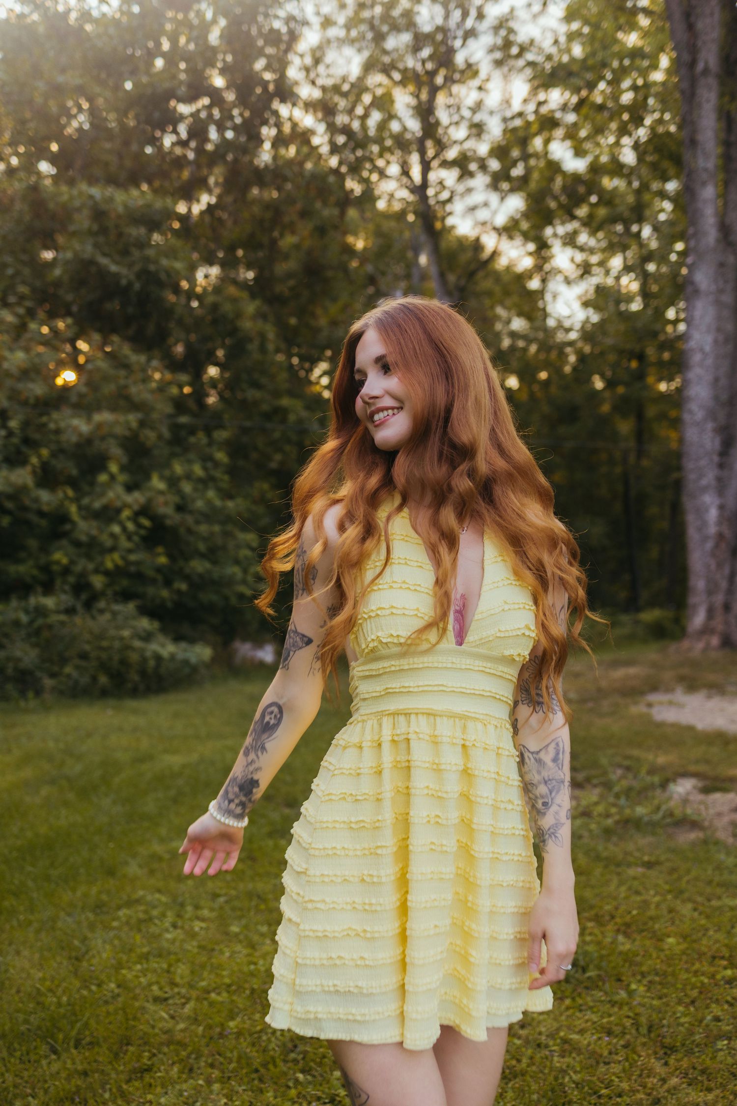 Smiling redhead woman with tattoos wearing a yellow mini dress standing outdoors in a lush green park at sunset.