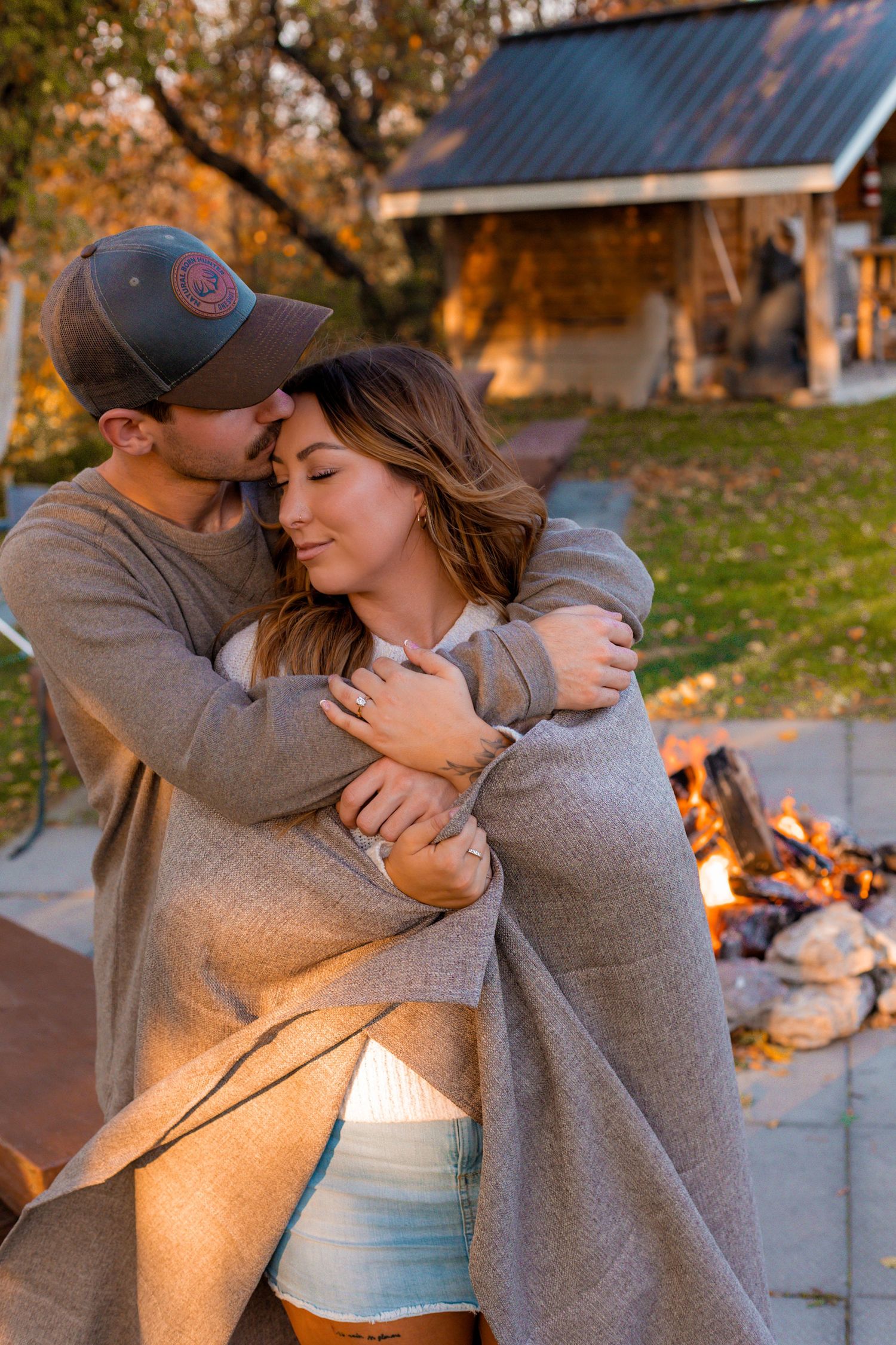 Couple wrapped in a cozy blanket embracing warmly near a glowing outdoor fire at sunset.