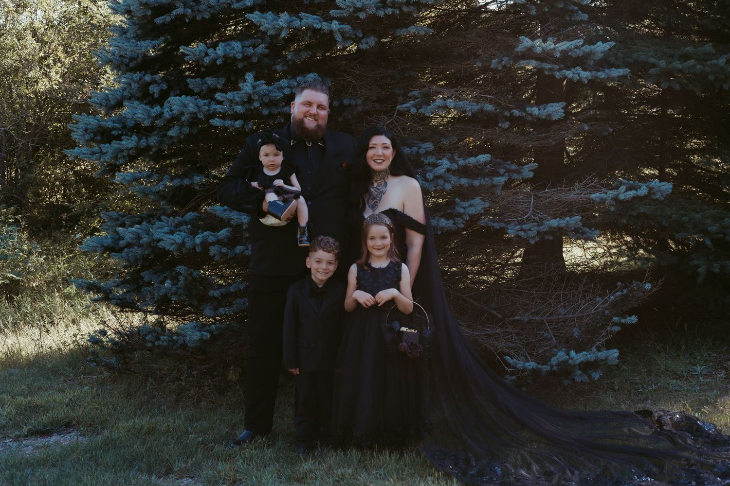 Family of five dressed in black posing outdoors near a blue spruce tree.