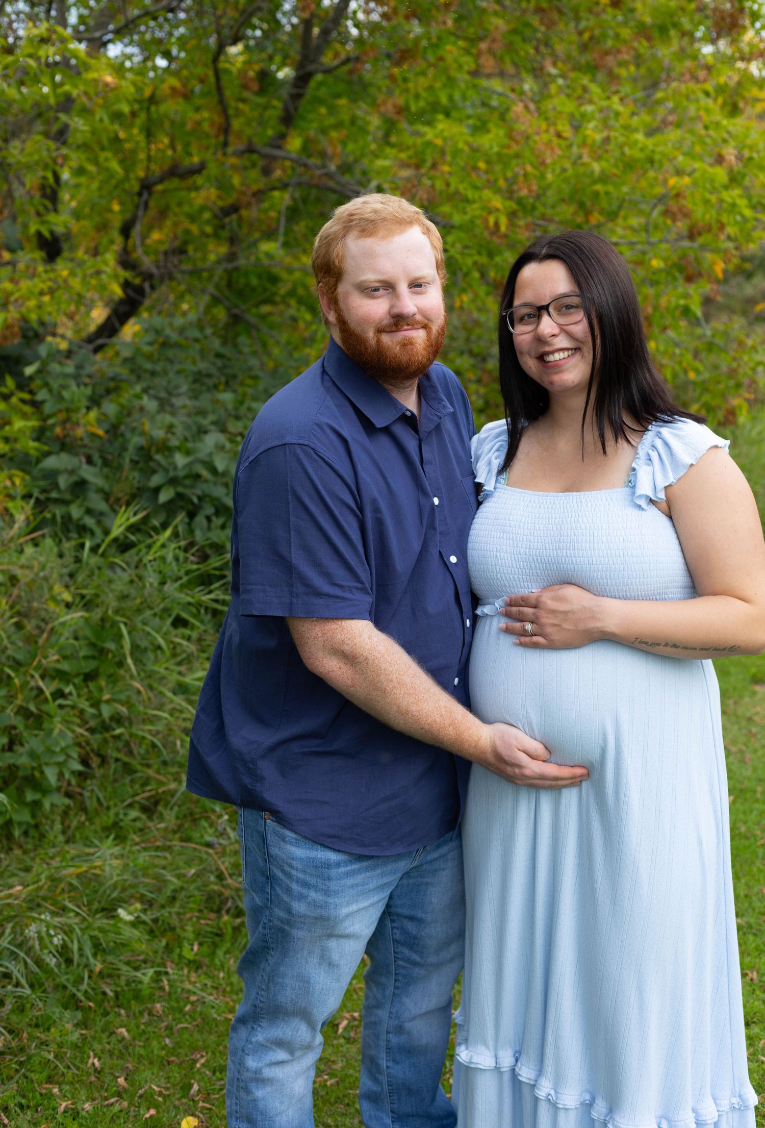 Smiling pregnant couple posing outdoors, woman in light blue maternity dress with partner in navy shirt.