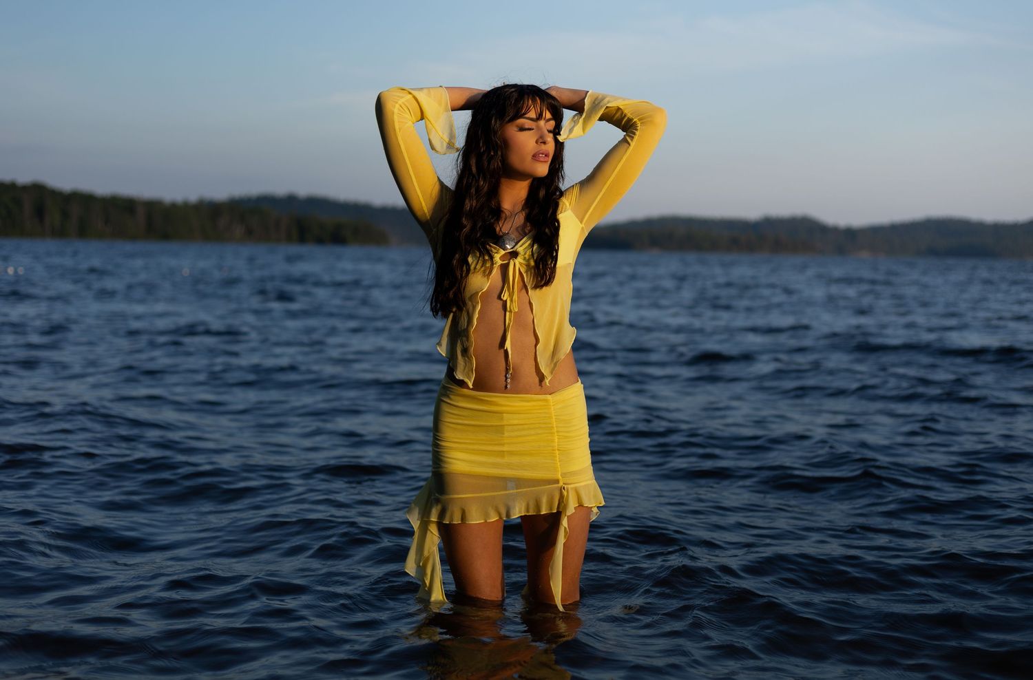 A woman in a yellow outfit stands confidently in a lake with mountains visible in the background.