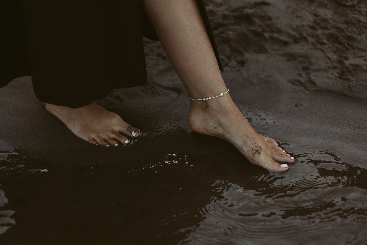 Bare feet with anklet jewelry standing in dark reflective water with small tattoo visible.