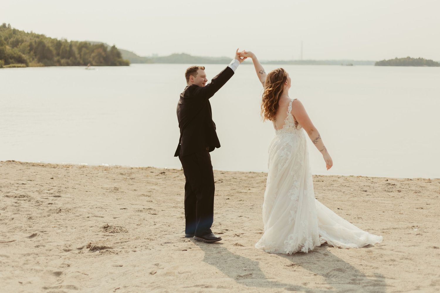 Groom twirls bride in white lace wedding gown on sandy beach by water.