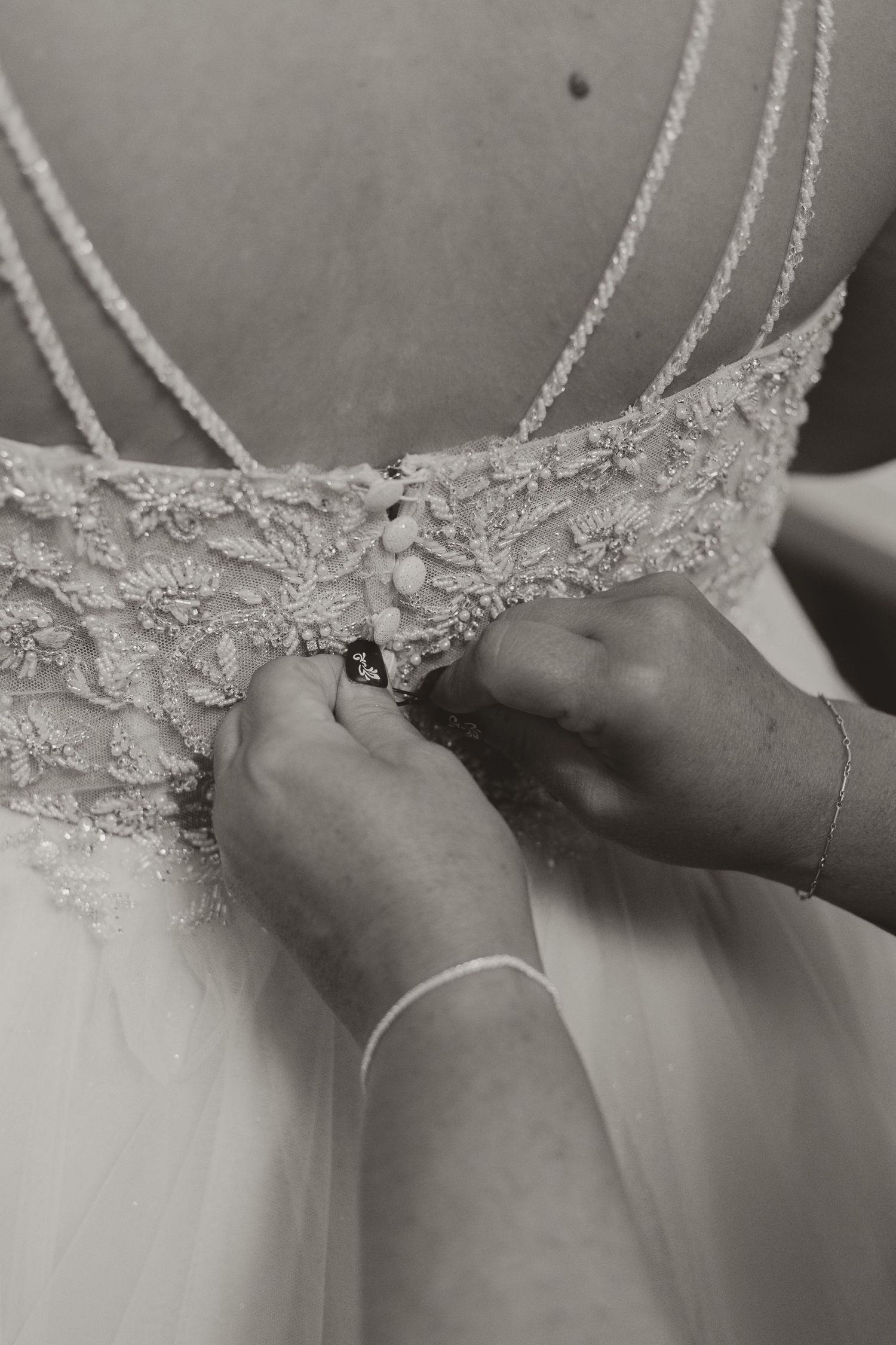 Black and white close-up of hands fastening delicate lace buttons on back of bridal gown.
