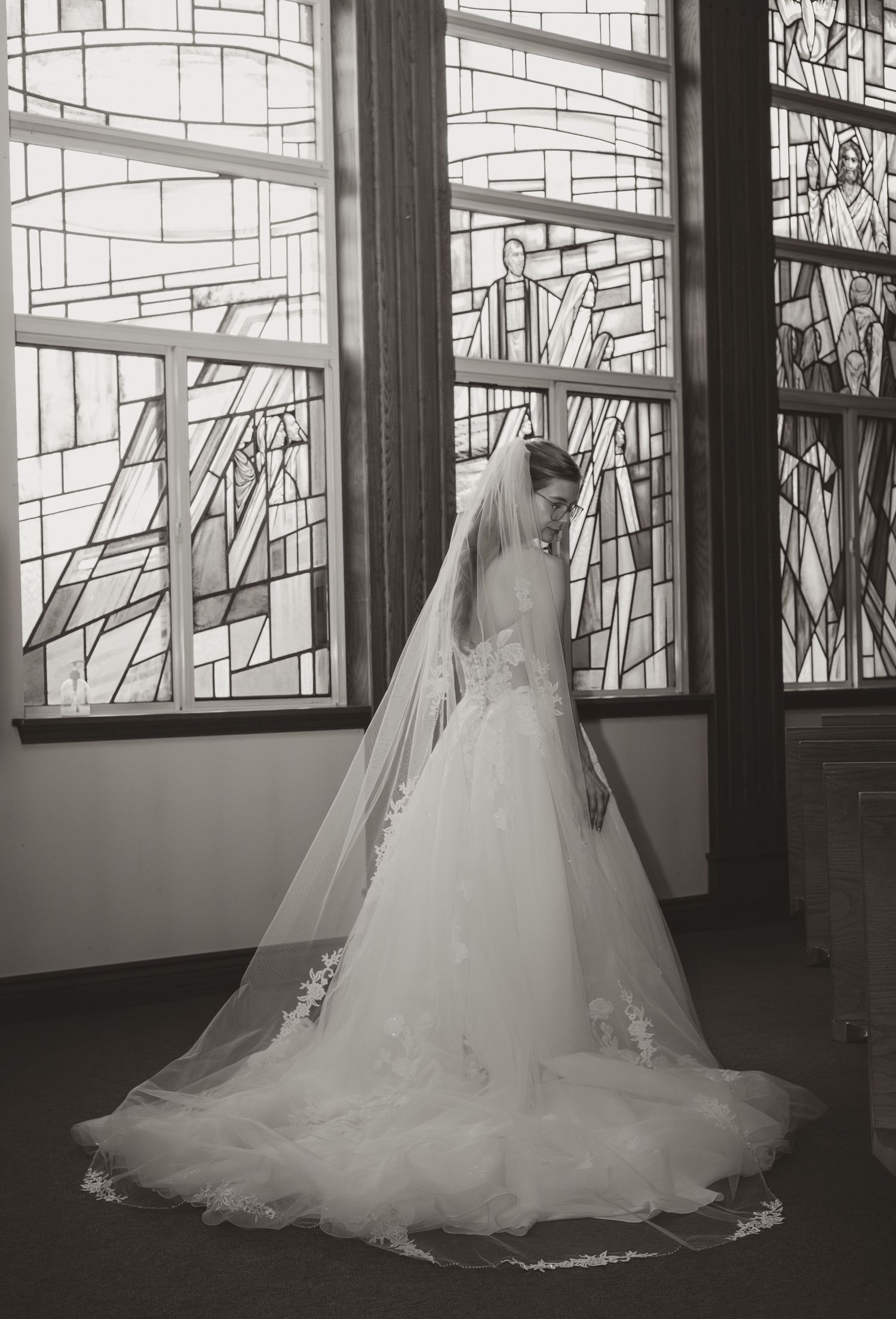 Elegant bride in a long lace wedding gown and veil standing before stunning stained glass church windows.