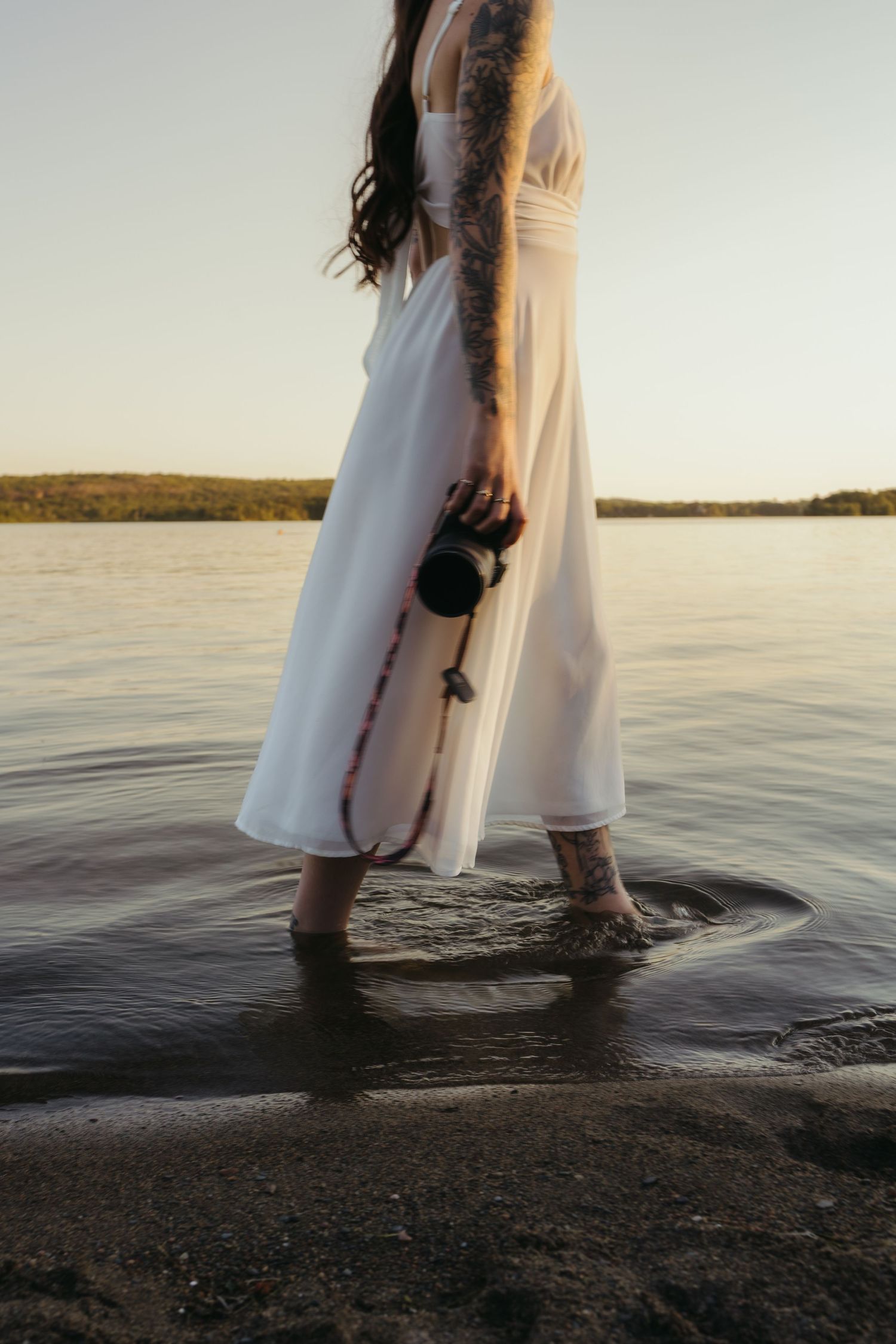 Tattooed woman in white dress wading in shallow lake water while holding a camera at sunset.