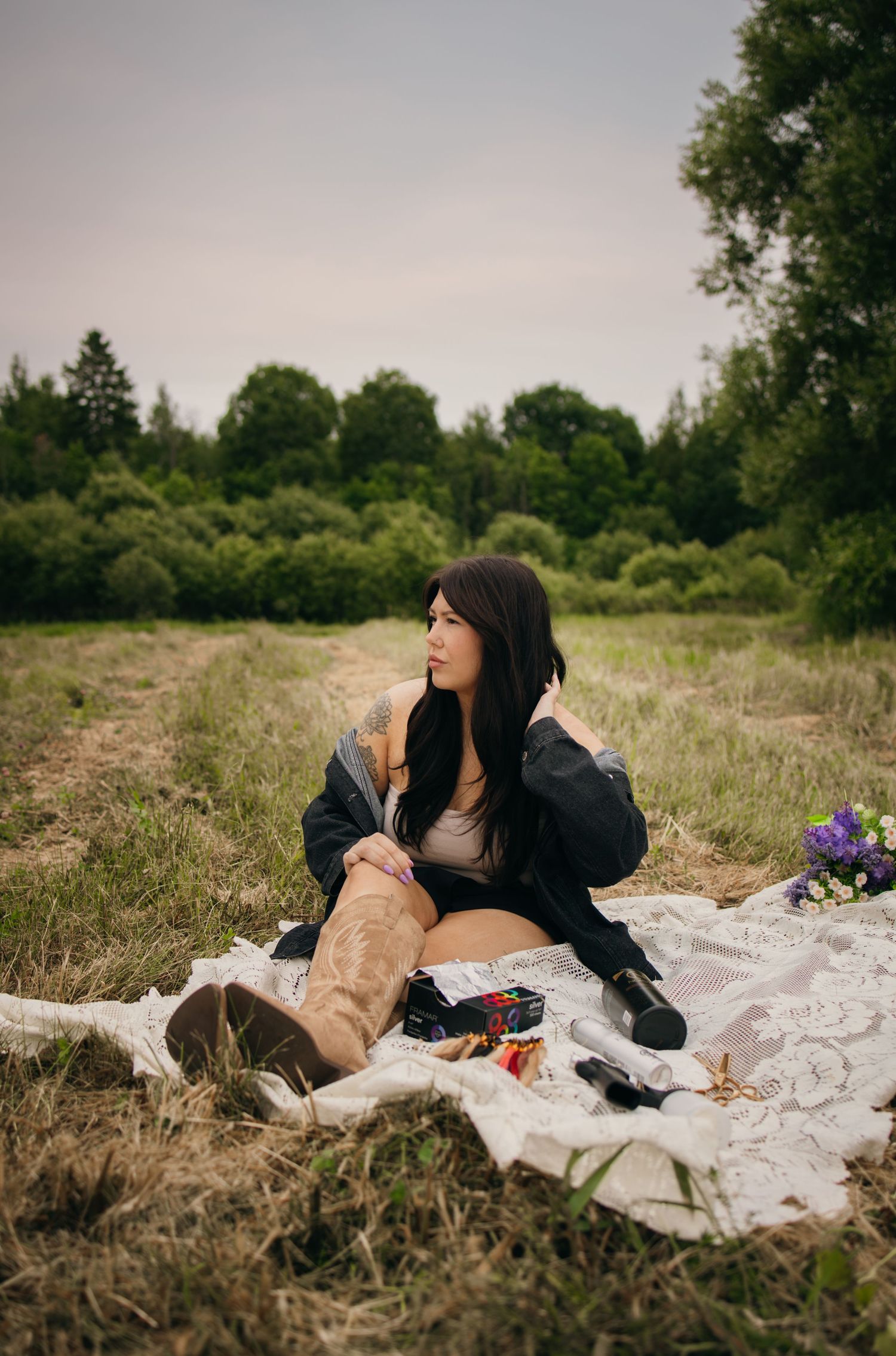 A dark-haired woman in a black jacket and brown boots sits on a lace blanket in a green meadow field.