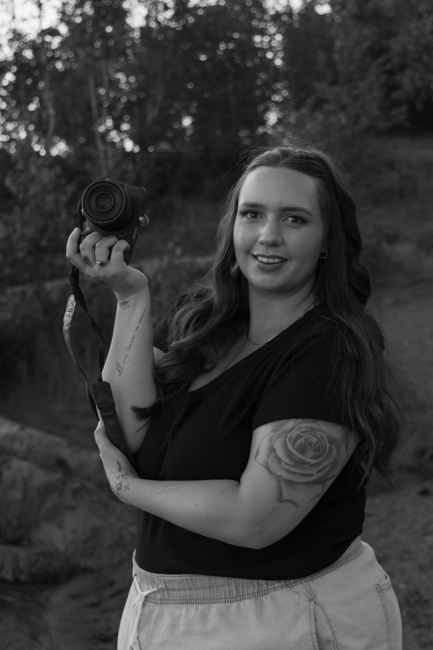 Black and white portrait of a young woman with long hair holding a camera, showcasing a rose tattoo on her arm.