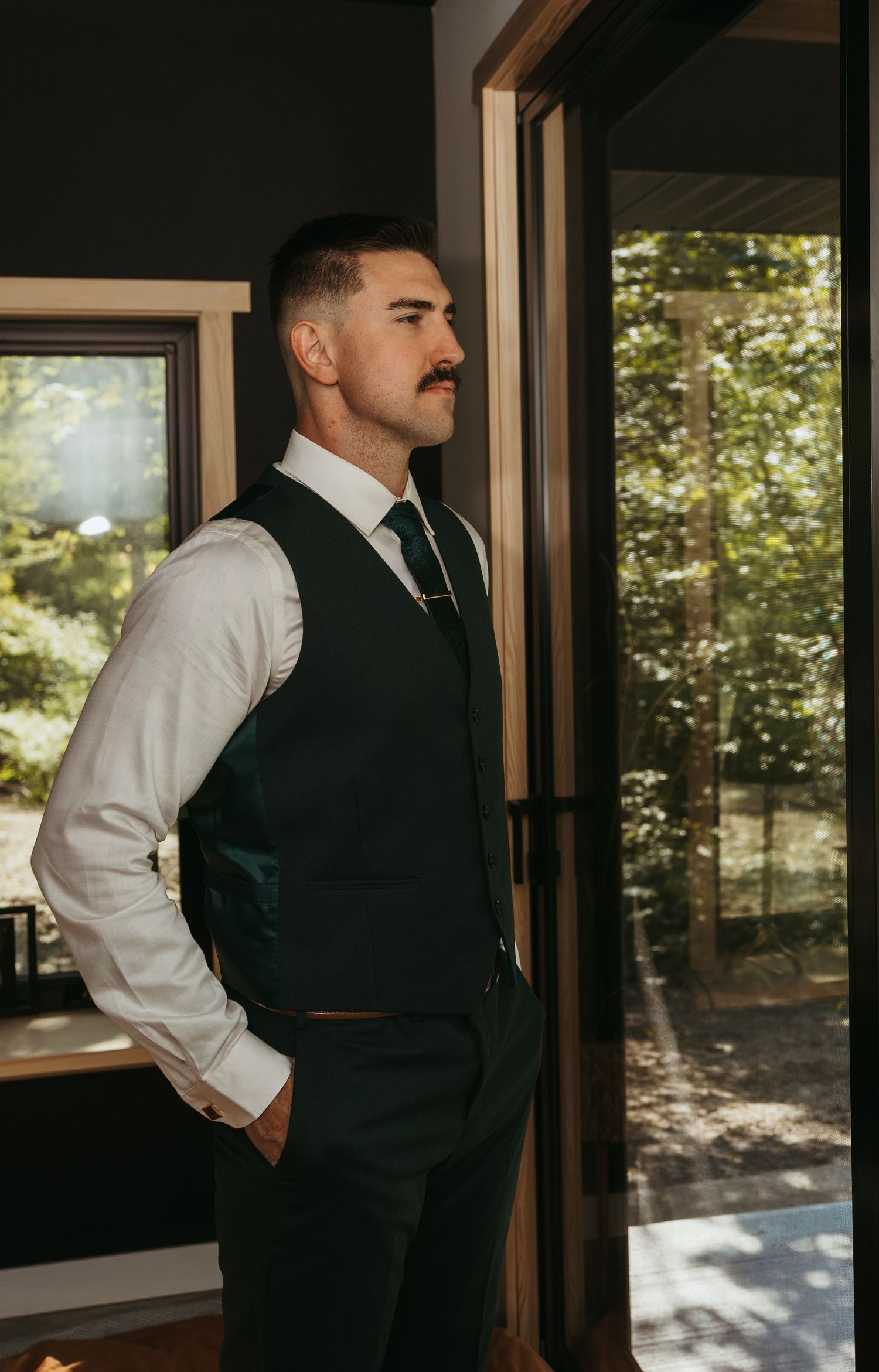 A dapper groom in a dark vest and tie gazes out a glass door surrounded by lush green trees on his wedding day.