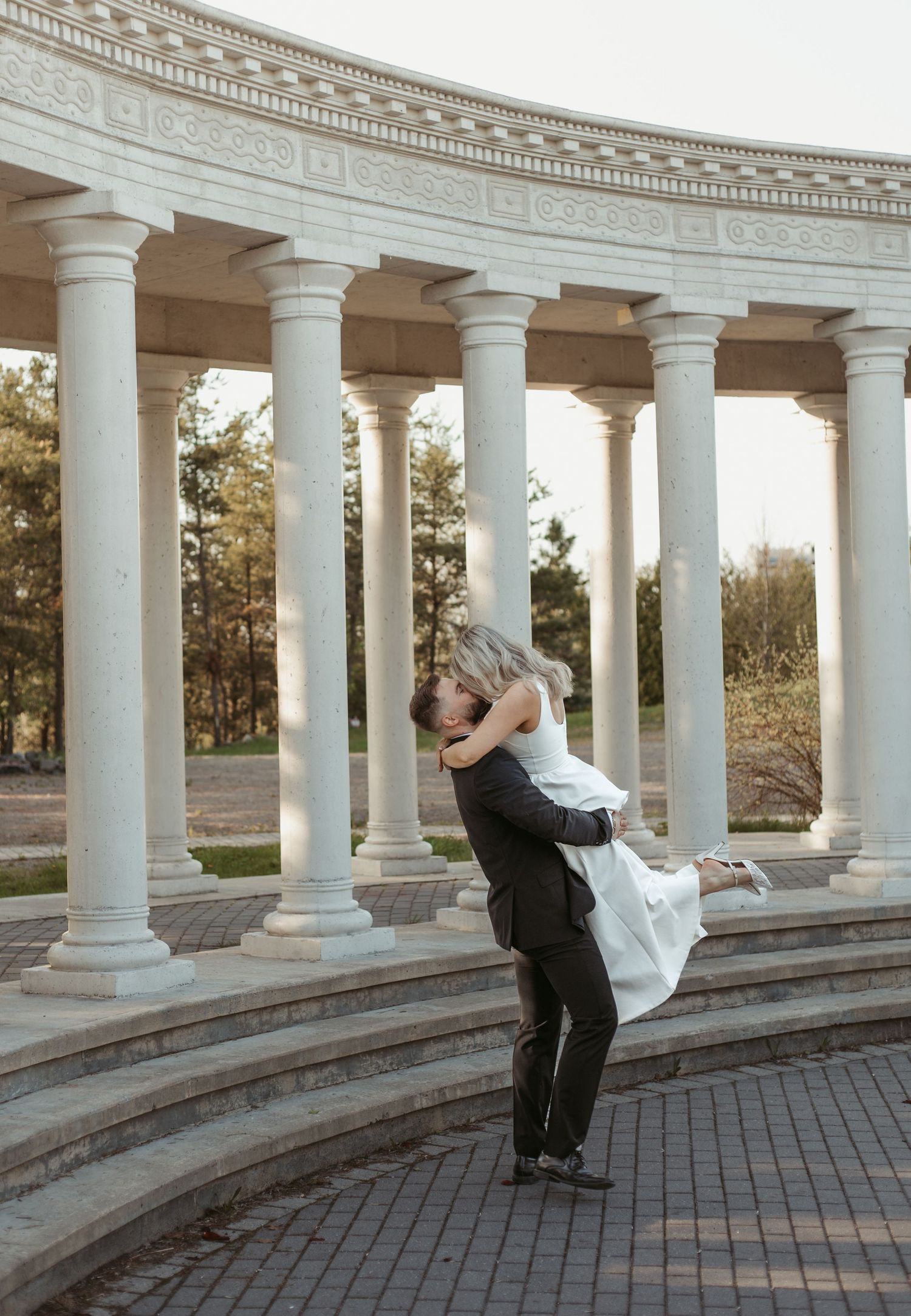 Groom lifting bride in white dress at elegant columned rotunda outdoor wedding photo.