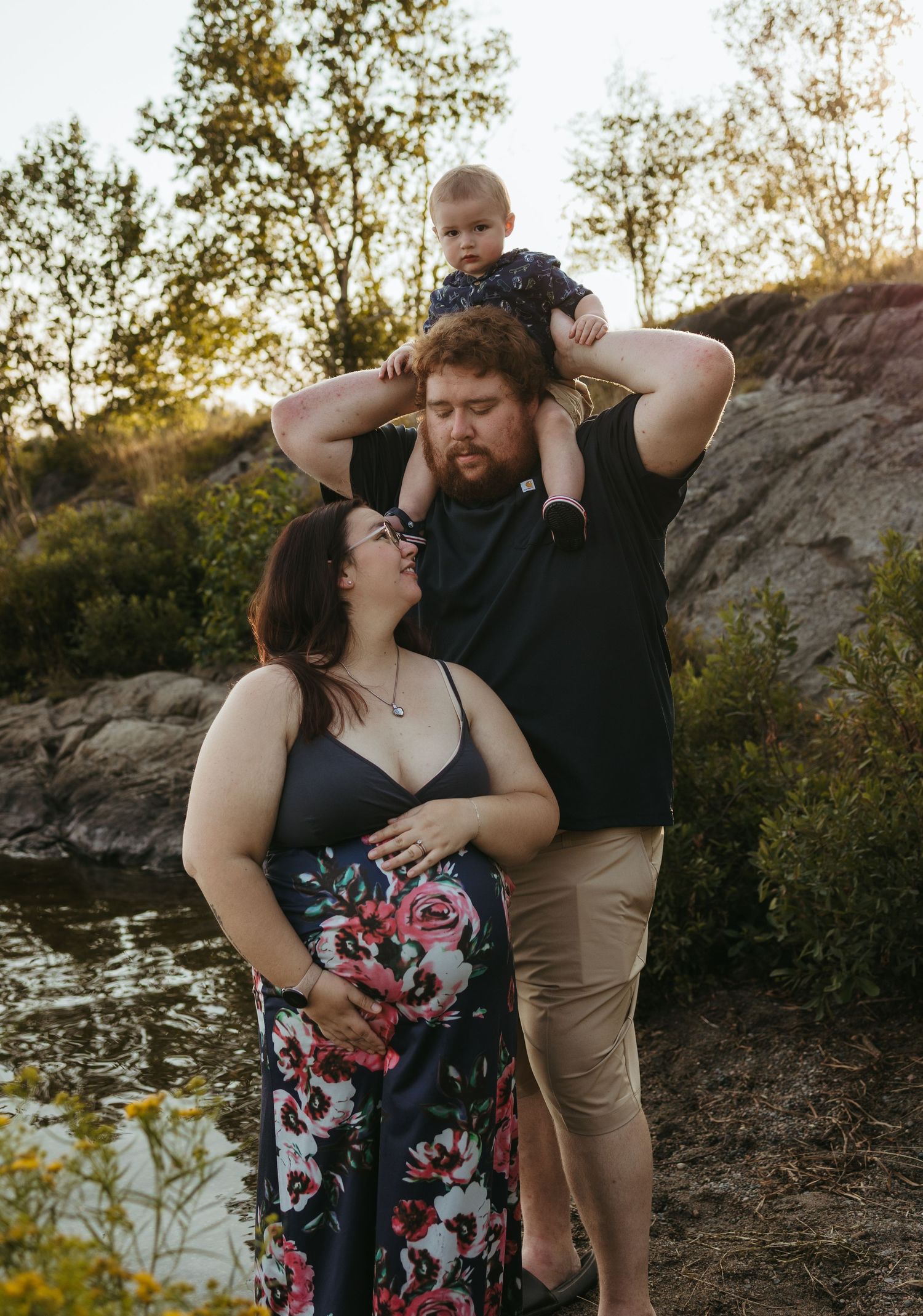 Pregnant woman in floral dress with partner holding toddler on shoulders outdoors near water at golden hour.