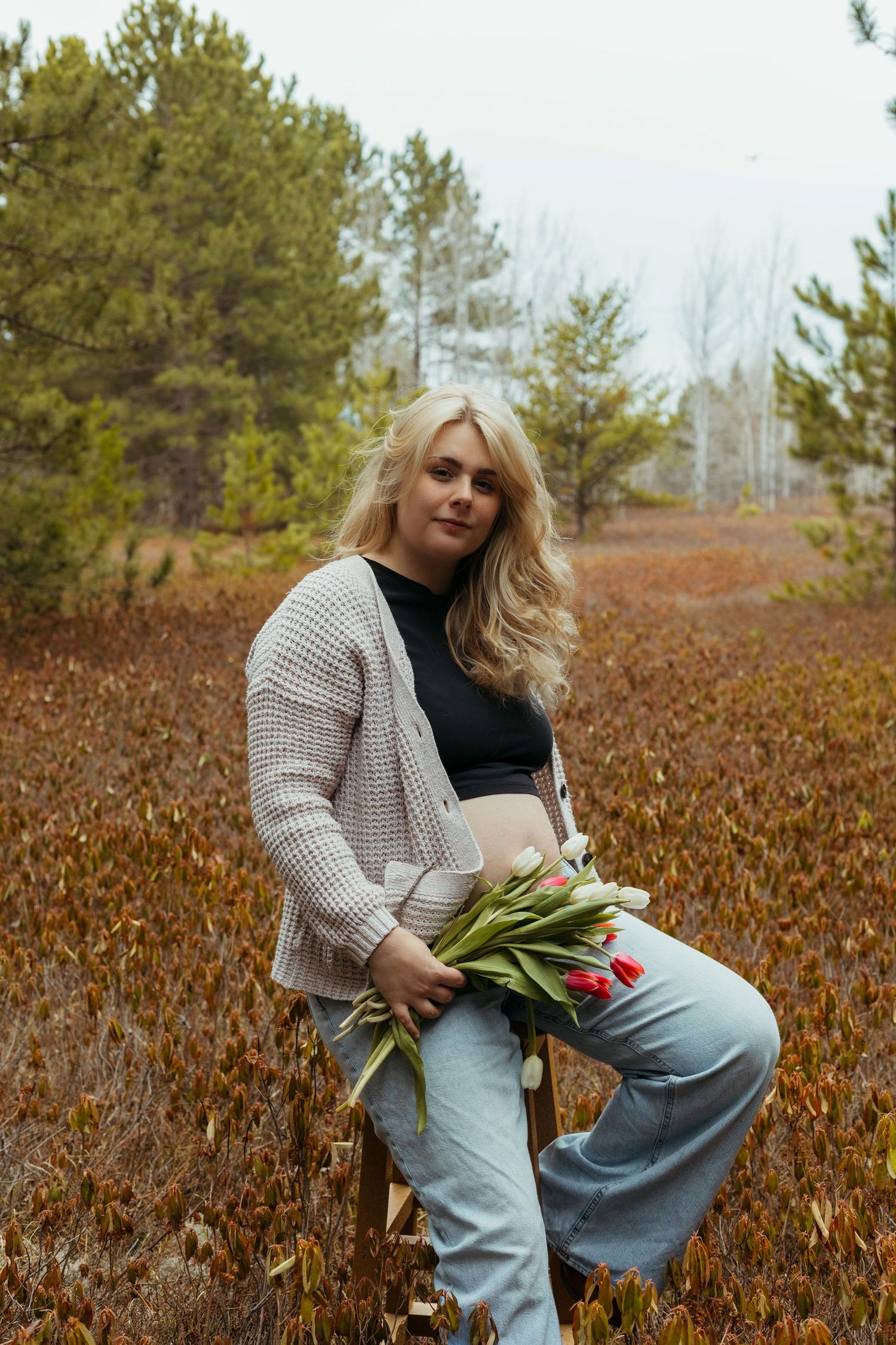 Blonde woman holding red tulips sits outdoors in a knit cardigan, crop top, and jeans surrounded by autumn foliage.