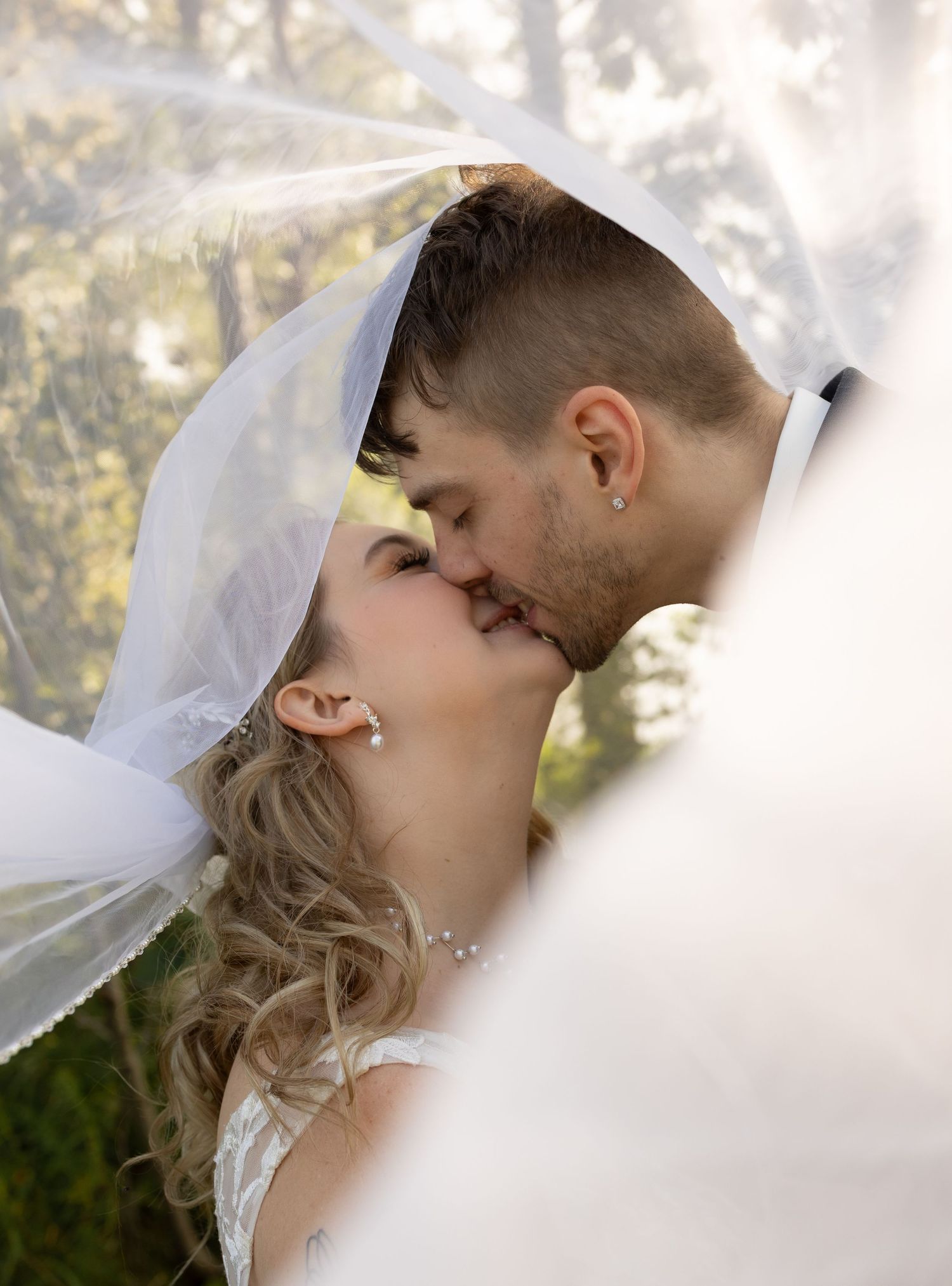 Bride and groom sharing a romantic kiss under a flowing white wedding veil outdoors.