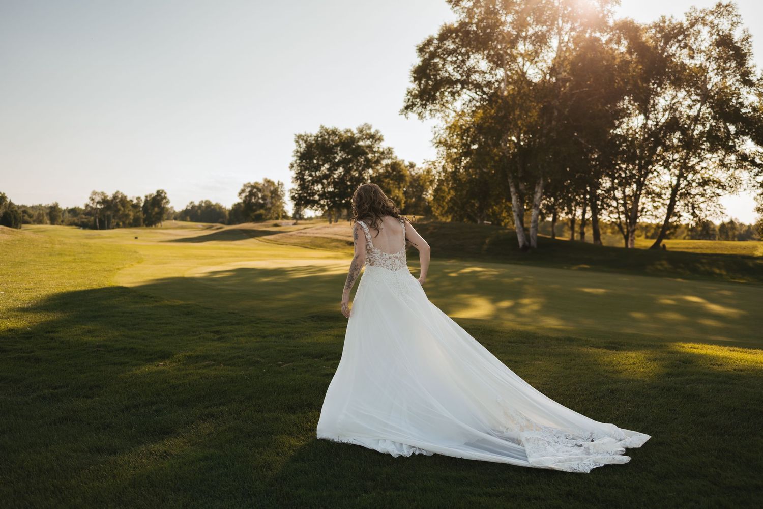Bride in elegant white wedding gown with long train walks across lush green golf course at golden sunset.