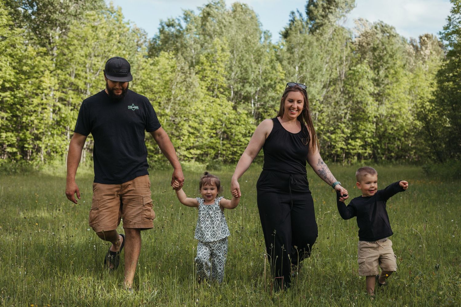 Family of four walking hand-in-hand through a green meadow surrounded by lush trees on a sunny day.