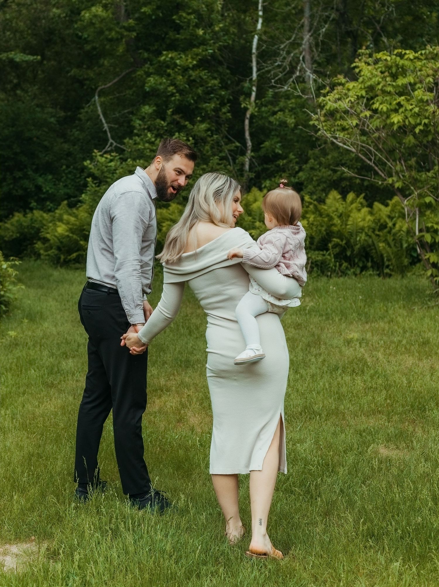 A young family poses outdoors on green grass, man in gray shirt holding hands with woman in white dress holding a baby.