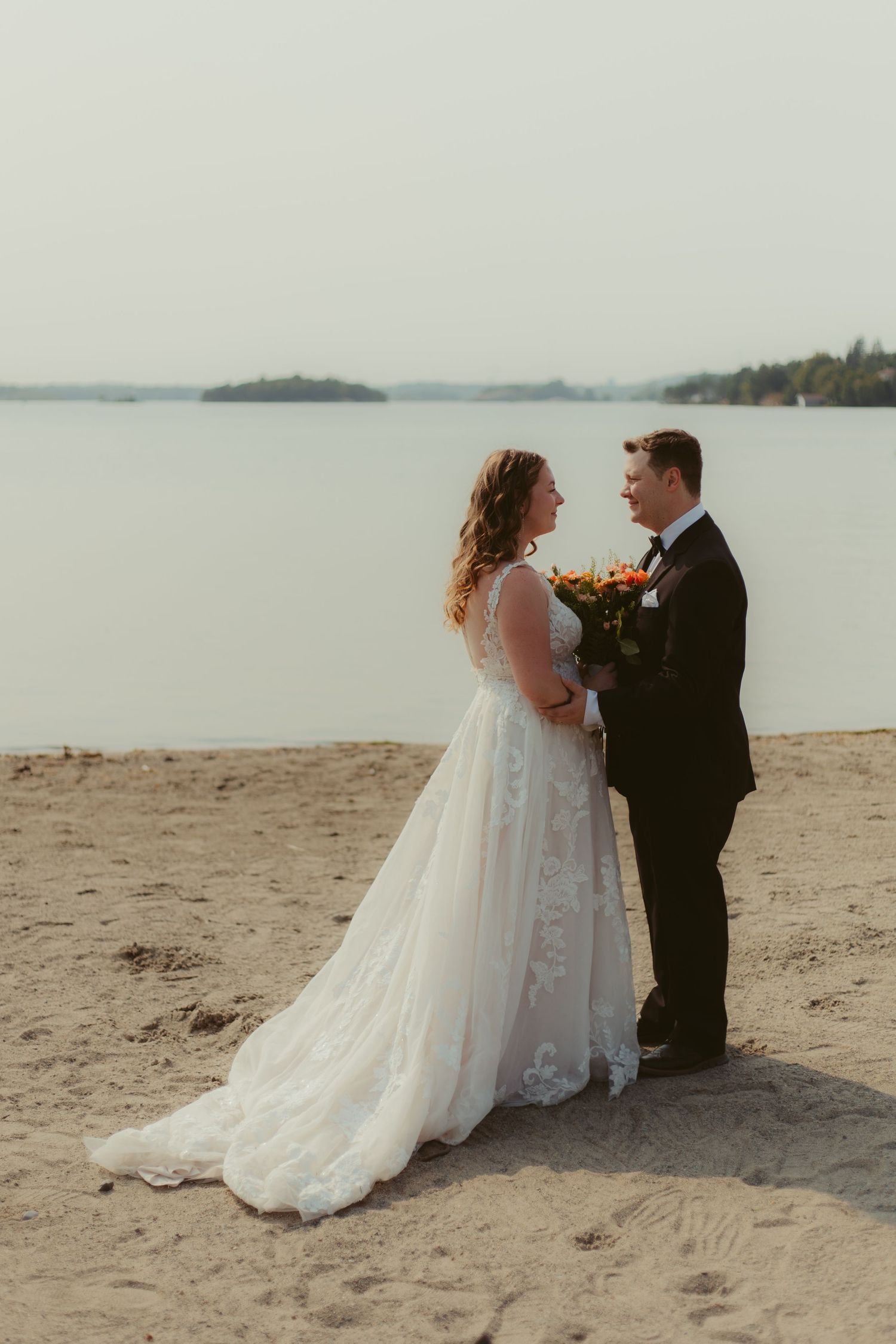 Bride and groom standing on sandy beach by calm lake, bride in lace gown holding bouquet, groom in black tuxedo.