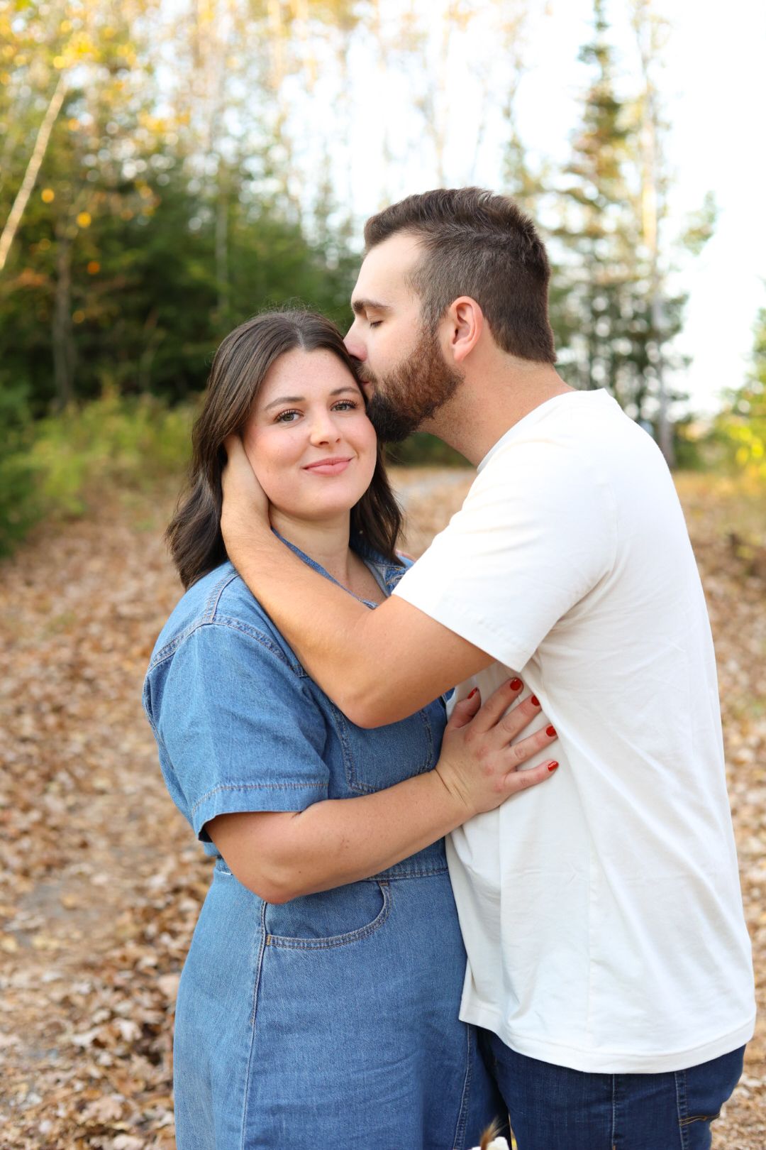 Man in white shirt tenderly kisses woman in denim dress on a leaf-covered outdoor path in fall.