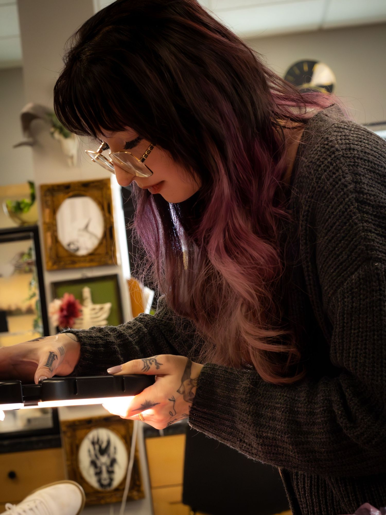 Woman with purple-tinted hair and tattoos working at a tattoo studio counter.