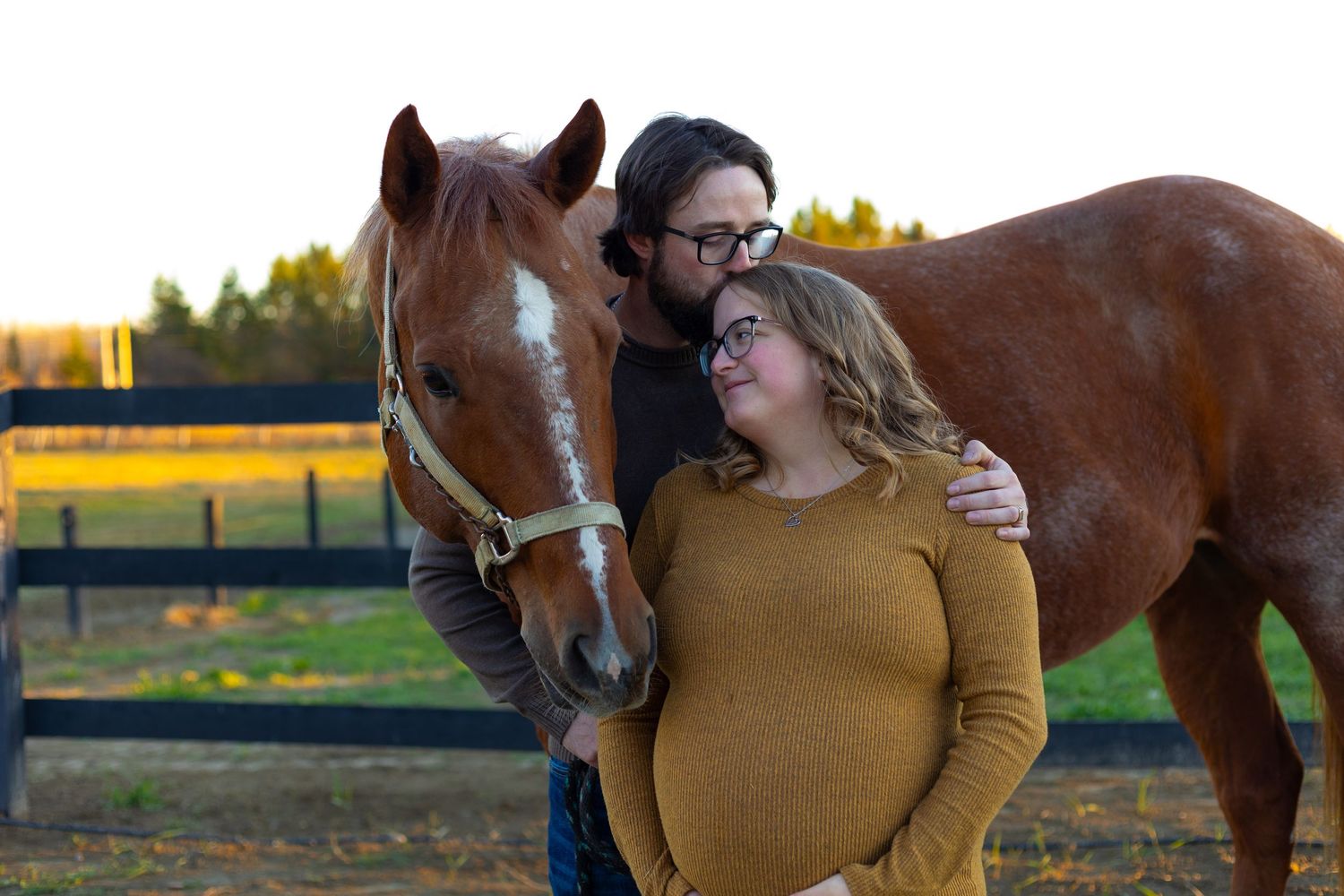Couple with pregnant woman posing beside chestnut horse at farm during golden hour.