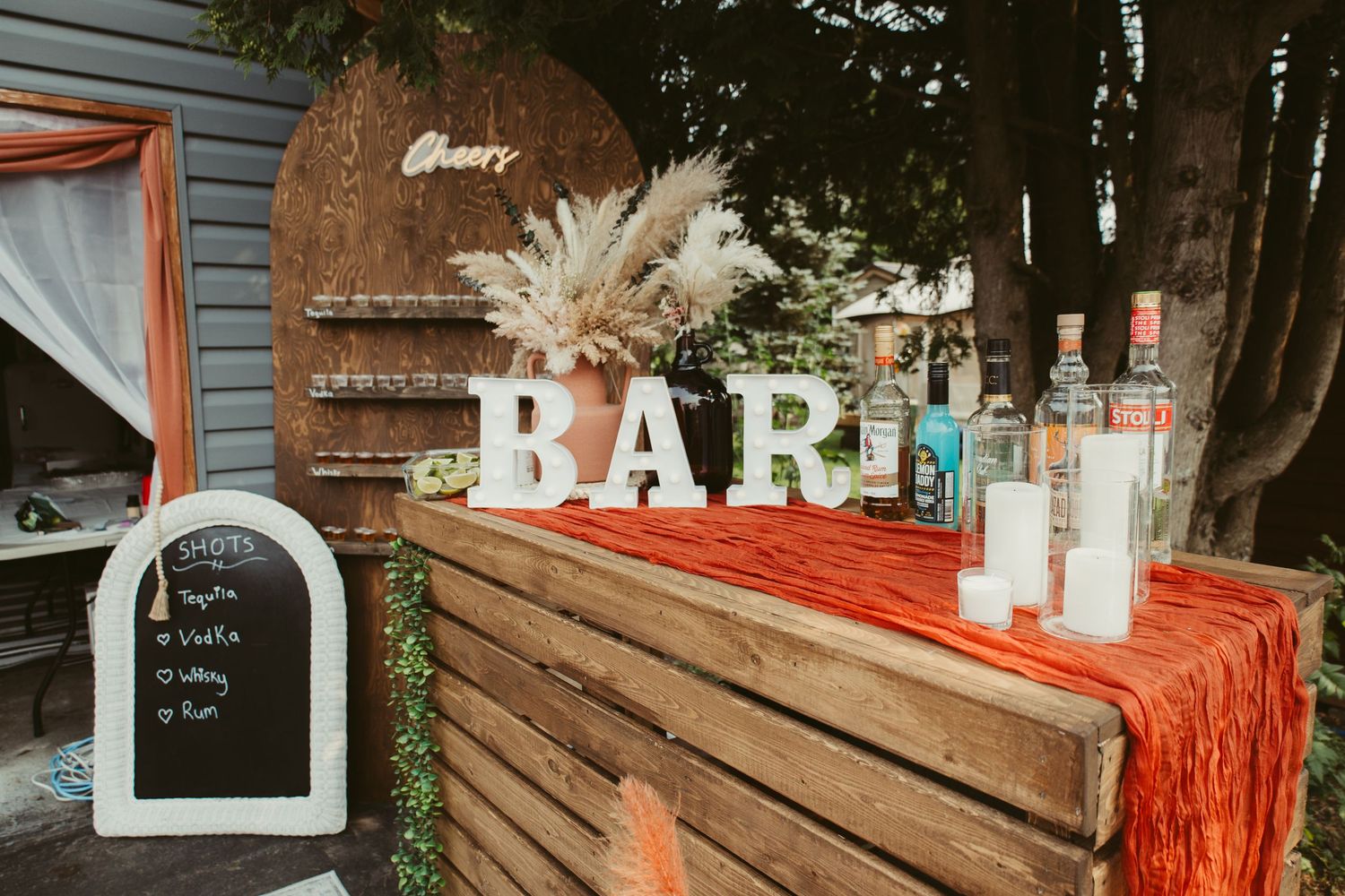 Rustic outdoor wedding bar setup with orange runner, liquor bottles, and BAR sign.