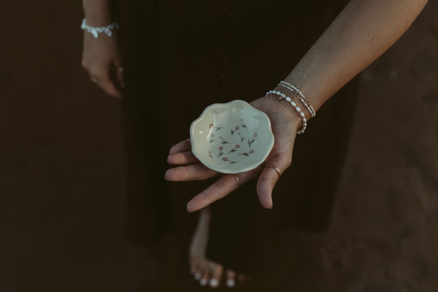 Hand holding a small white ceramic dish with delicate fish design, wearing stacked bracelets.