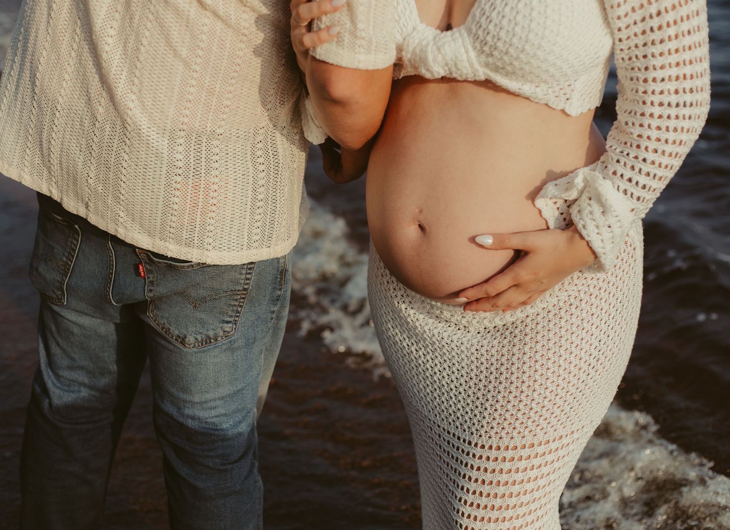Close-up of pregnant couple at beach, partner embracing woman's baby bump near ocean waves.