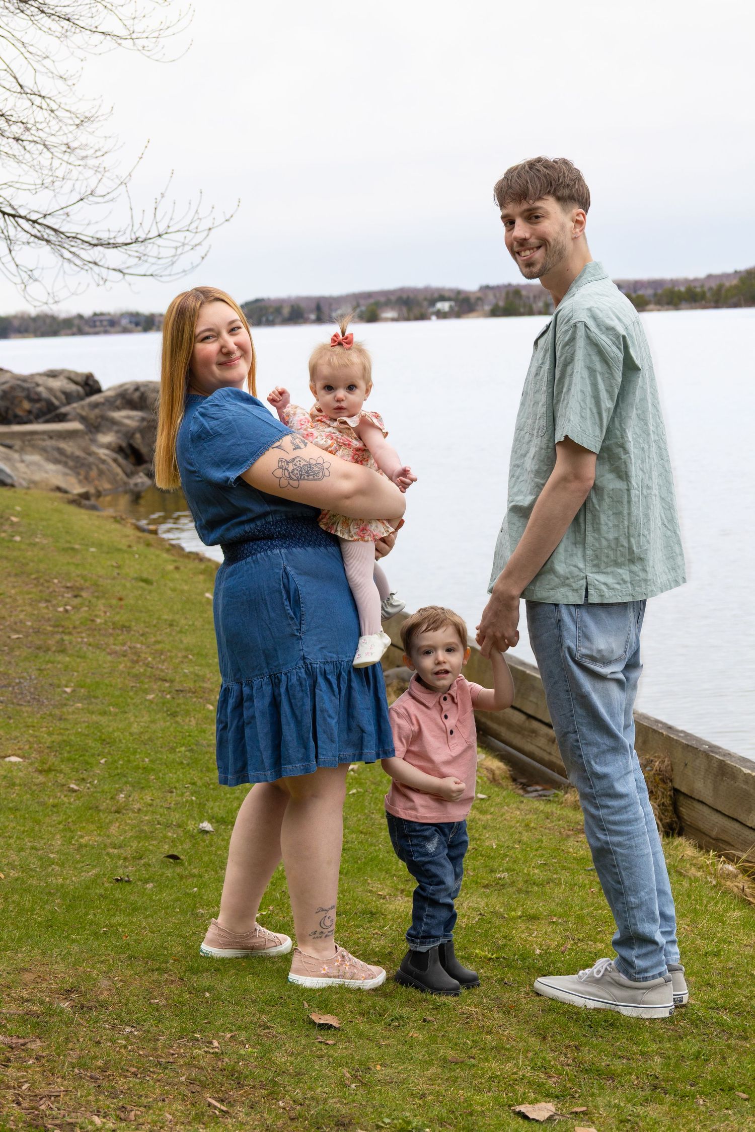 Family of four posing outdoors near a lake, mother holding baby girl, father smiling, toddler standing between them.