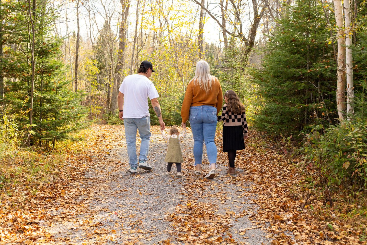 Family of four walks hand-in-hand down a leaf-covered forest path in fall.