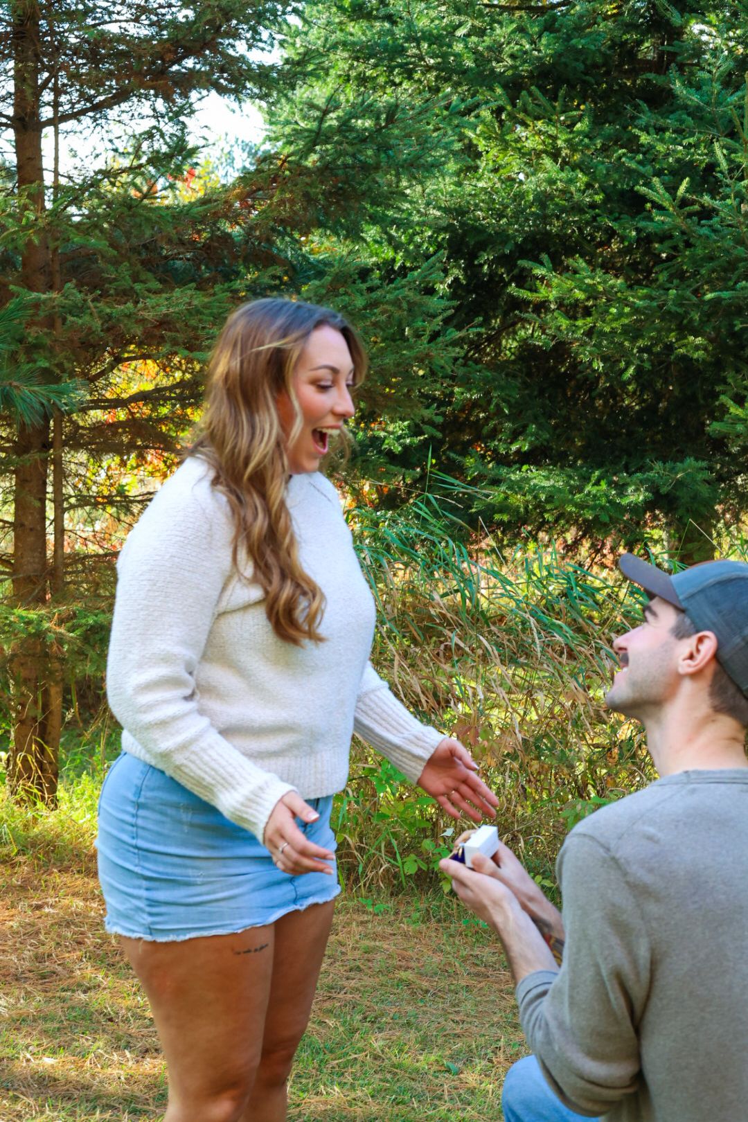 Man proposing on one knee to surprised woman in forest clearing.