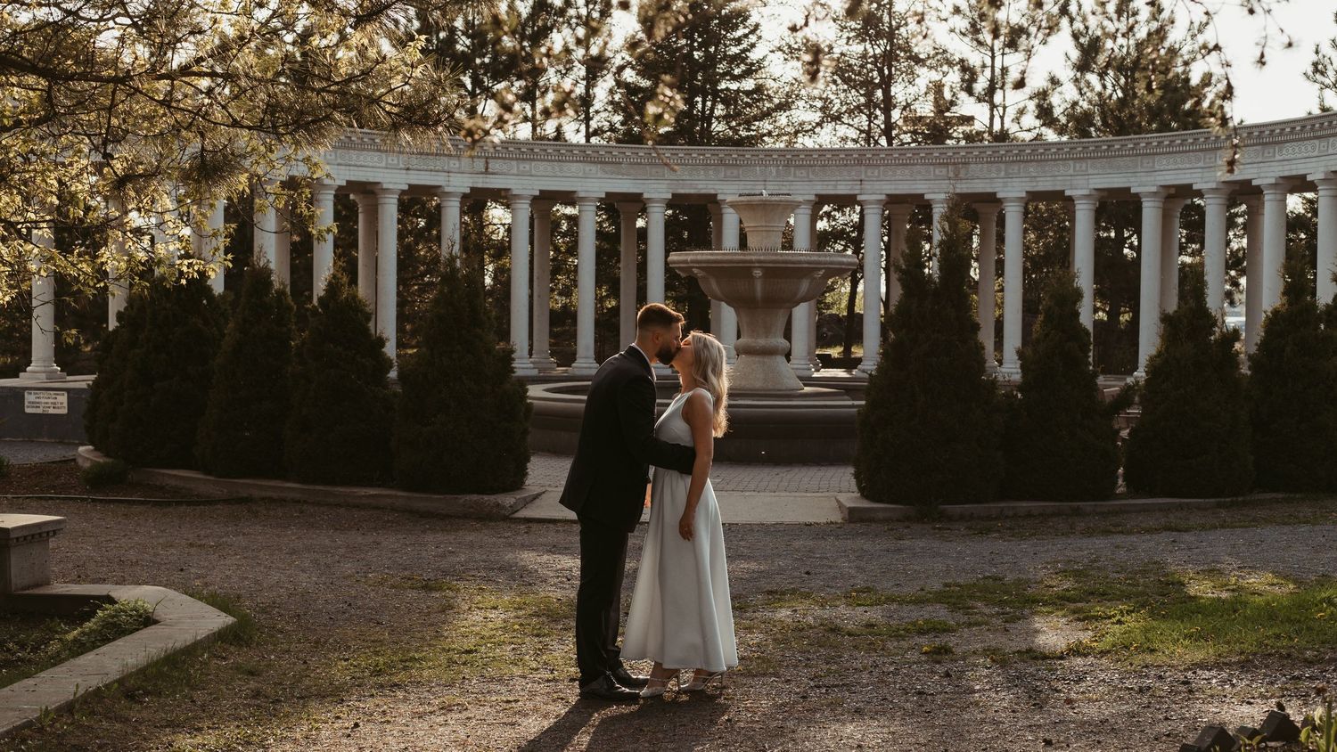 A bride and groom share a romantic embrace in a garden with white columns, evergreen trees, and a fountain backdrop.