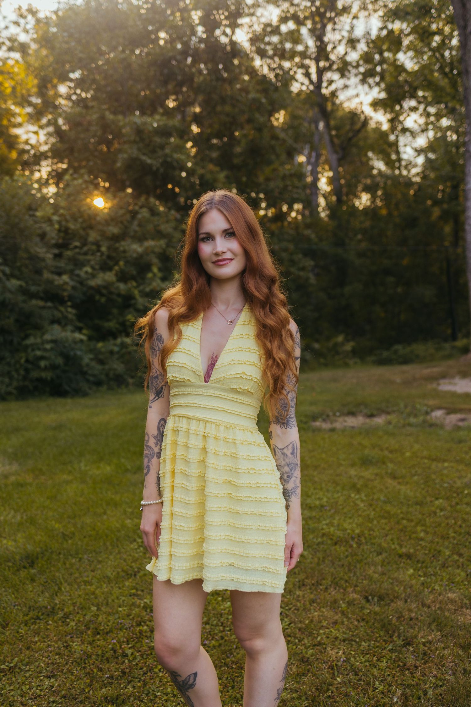 Young woman with red hair and tattoos wearing a yellow sundress standing in a green park at golden hour sunset.