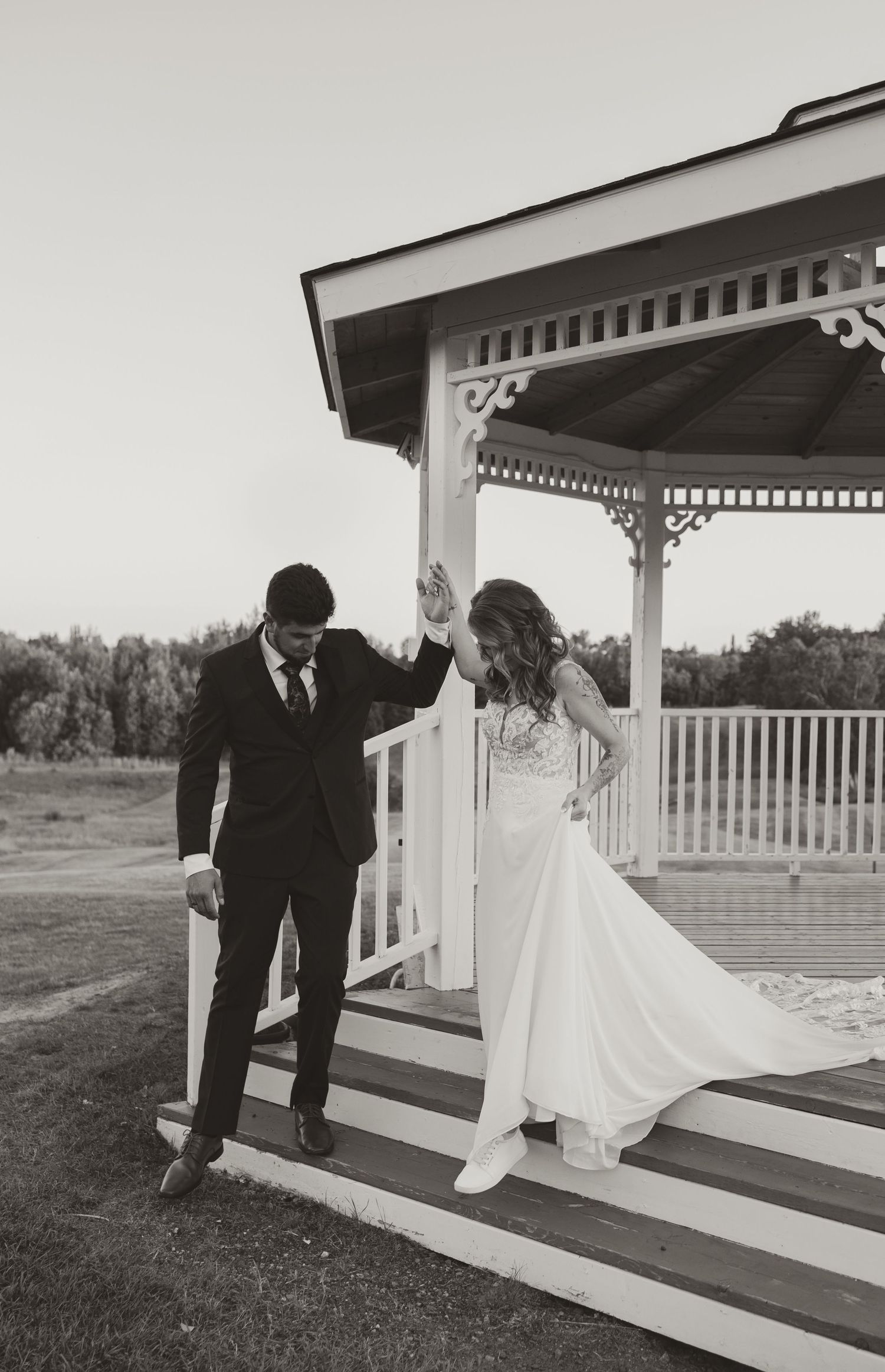 Black and white photo of a bride and groom holding hands on gazebo steps at their outdoor wedding.