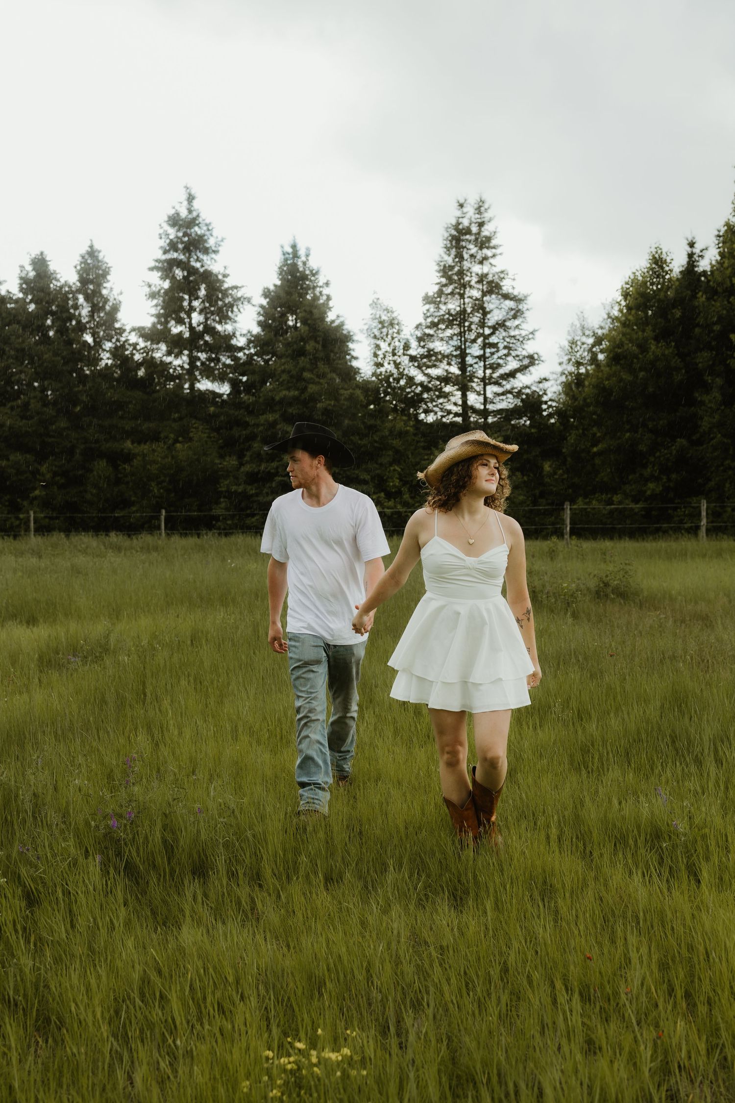 Couple holding hands walking through a lush green field with evergreen trees in background.