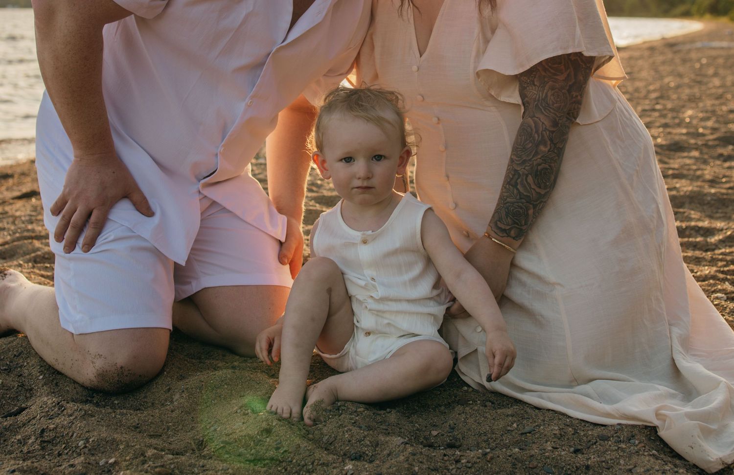 A toddler sits on sandy ground between two adults in pastel outfits during a warm outdoor family photo session.