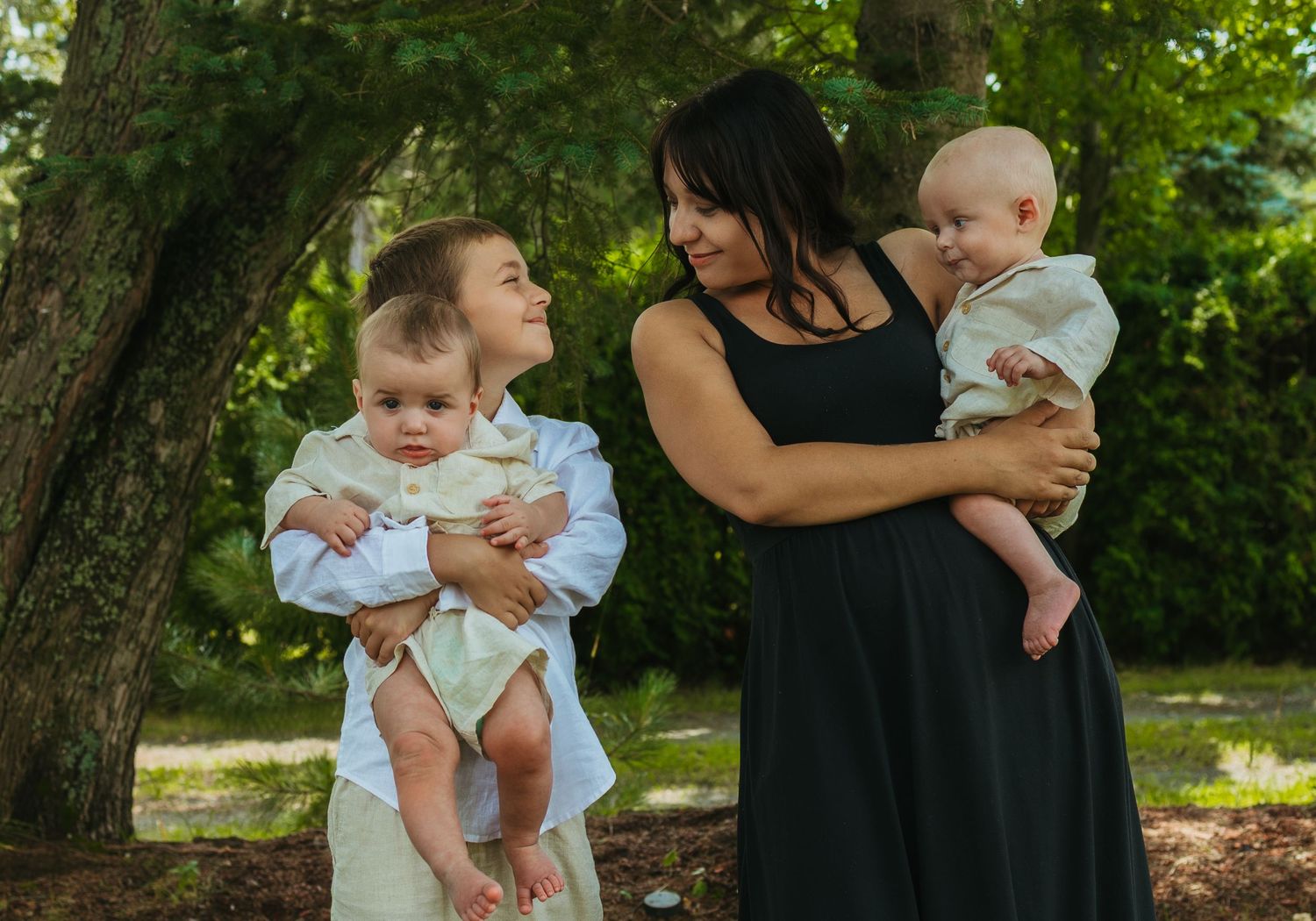 A woman in a black dress holds a baby outdoors while a child holds another infant near green trees.