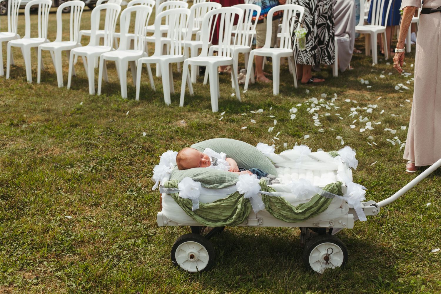 A sleeping baby in a decorated wagon at an outdoor wedding ceremony on green grass.