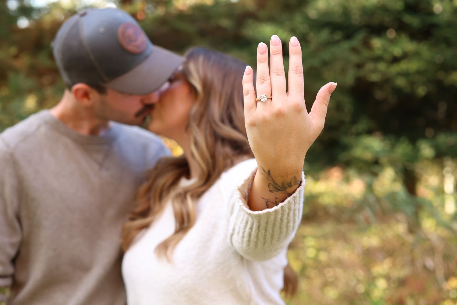 Newly engaged woman displays diamond ring while couple kisses in autumn setting.